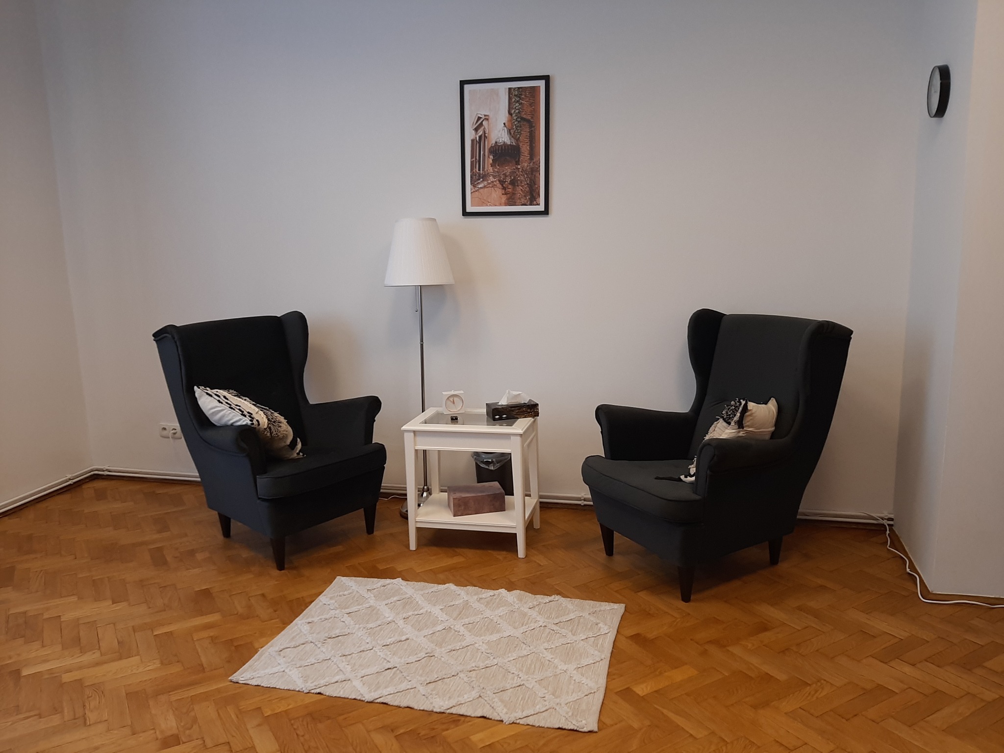 Pokój z dwoma grafitowymi fotelami typu uszak, białym stolikiem kawowym, lampą podłogową i dywanem z geometrycznym wzorem na parkiecie.