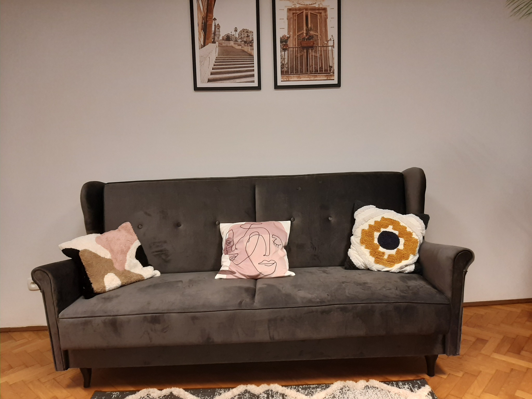 Szary welurowy fotel z trzema ozdobnymi poduszkami w pomieszczeniu z parkietem i dwoma obrazami na ścianie.