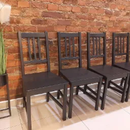 Rząd czterech ciemnych krzeseł z drewnianymi oparciami, ustawionych pod ceglaną ścianą, obok metalowego kwietnika z trawą pampasową.