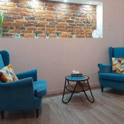 Gabinety Psychoterapii ARIADNA Katowice Raciborska - Jasne wnętrze gabinetu z dwoma turkusowymi fotelami z poduszkami w geometryczne wzory, stolikiem z chusteczkami higienicznymi i ceglaną ścianą z wnęką, w której ustawione są rośliny doniczkowe...