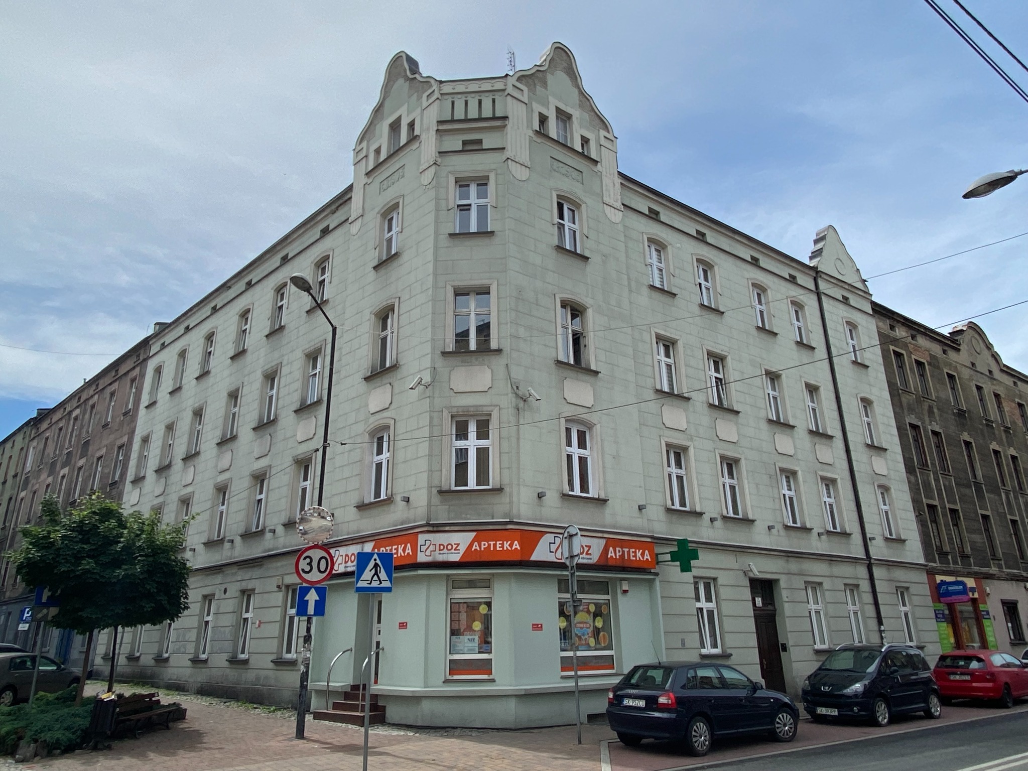 Fasada budynku z apteką DOZ na rogu, widoczne znaki drogowe i zaparkowane samochody.