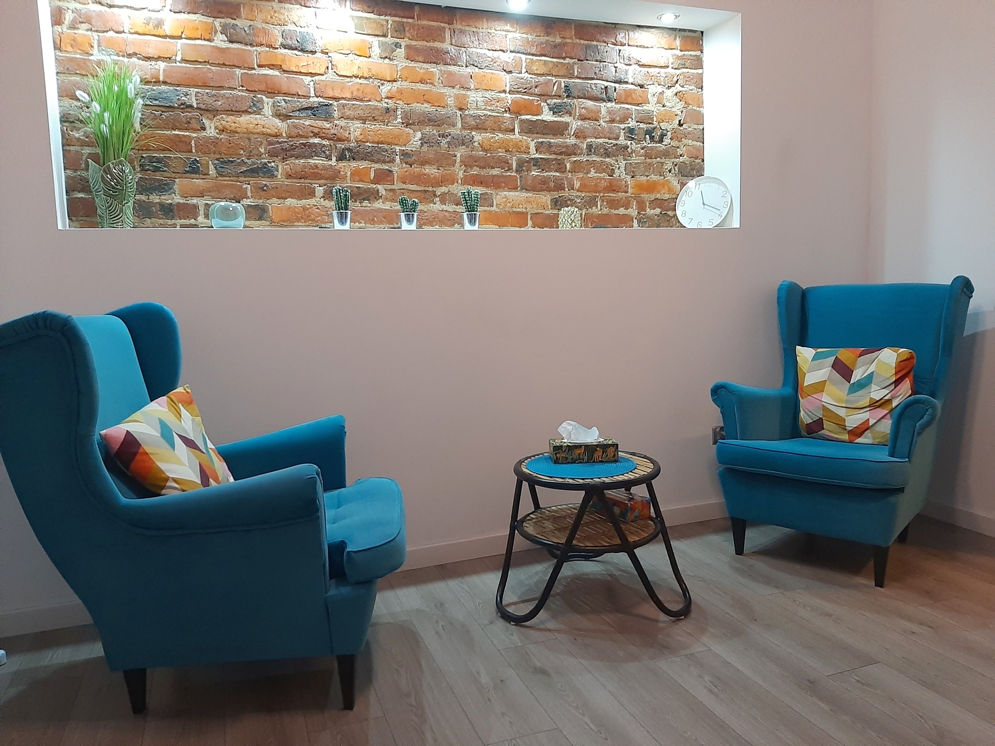 Jasne wnętrze gabinetu z dwoma turkusowymi fotelami z poduszkami w geometryczne wzory, stolikiem z chusteczkami higienicznymi i ceglaną ścianą z wnęką, w której ustawione są rośliny doniczkowe...