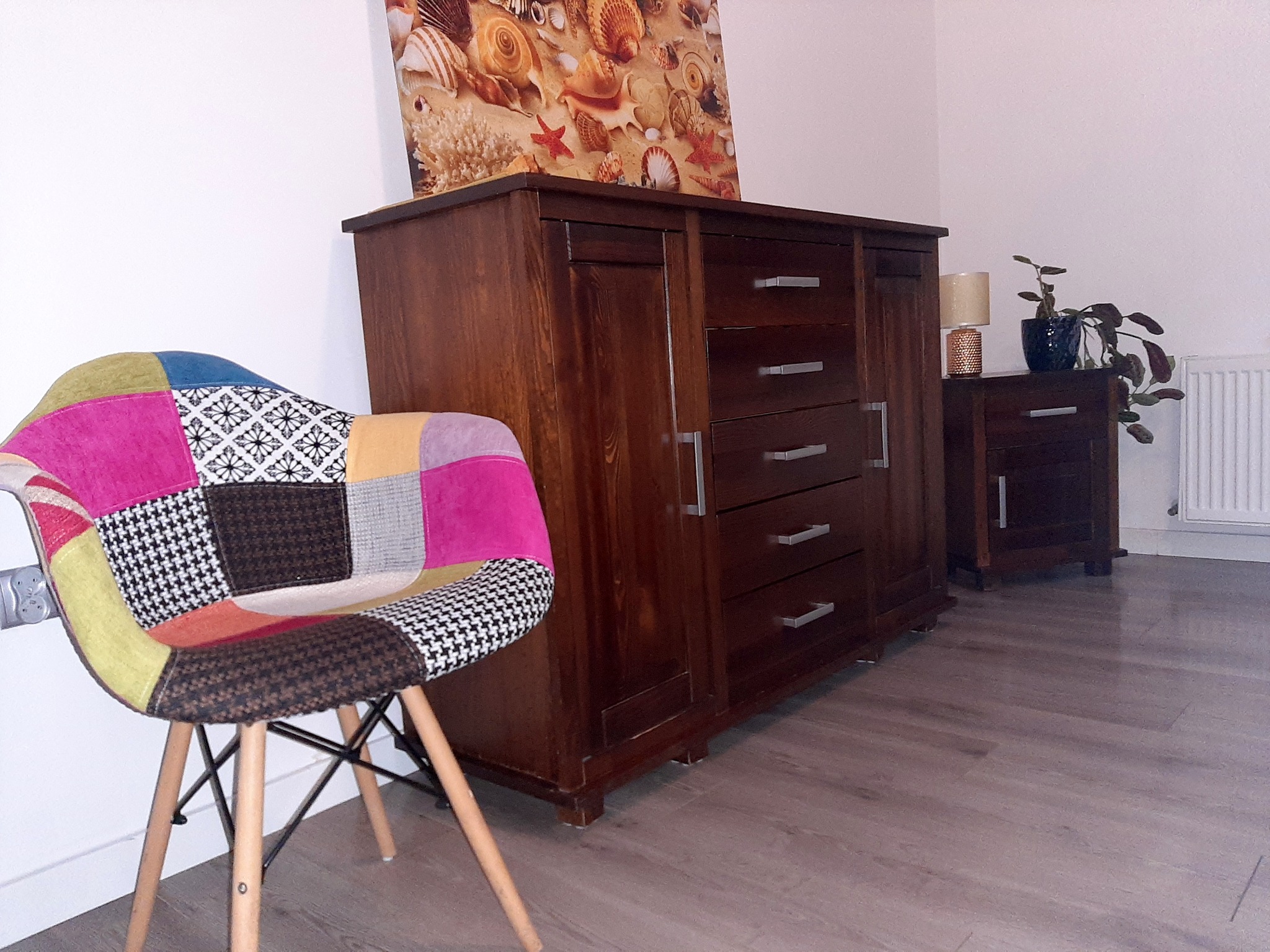 Pokój z drewnianą komodą, szafką nocną z lampką i rośliną doniczkową oraz patchworkowym krzesłem, nad komodą wisi obraz z motywem muszli.