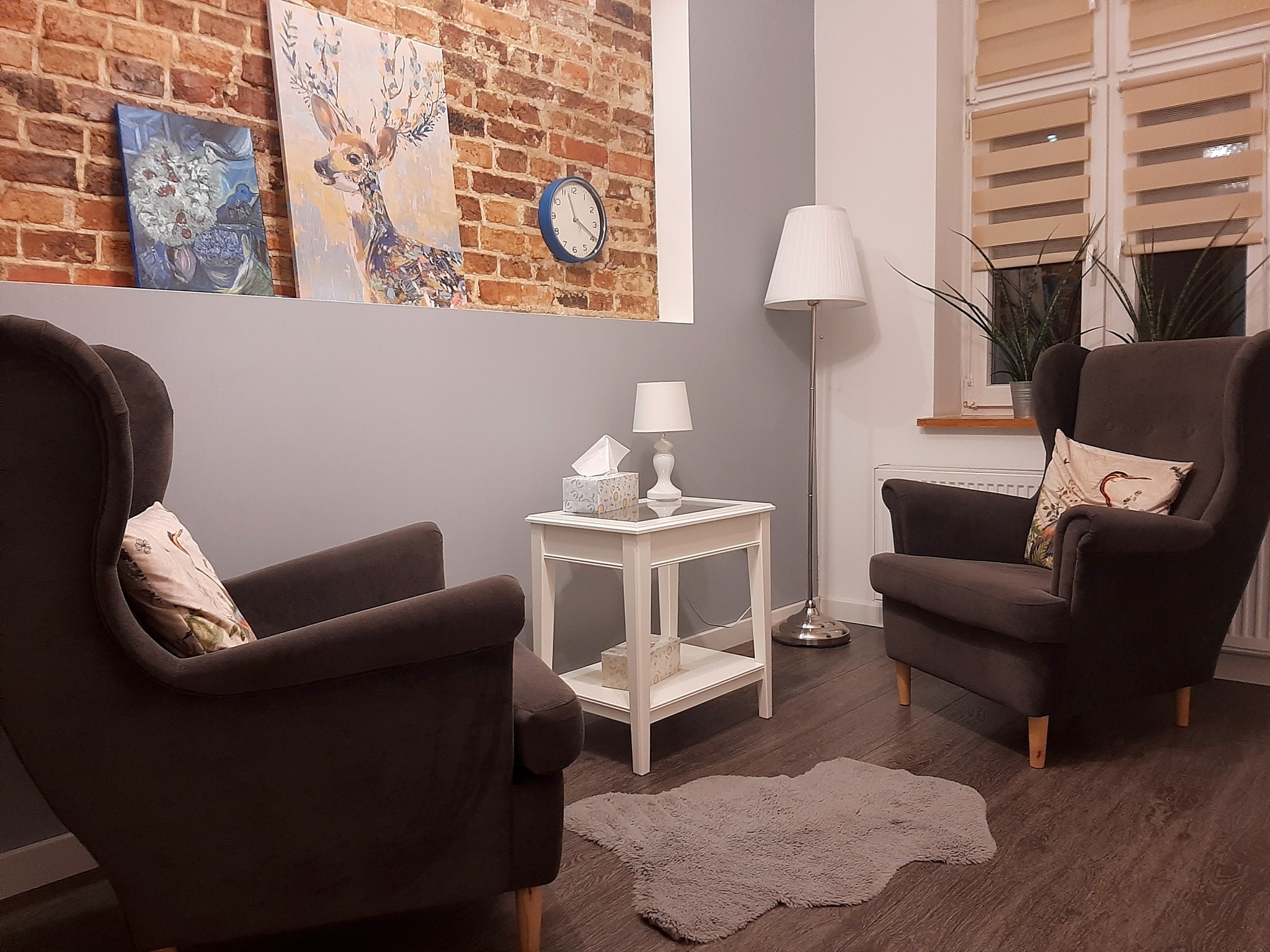 Przytulny gabinet z dwoma fotelami typu uszak, małym białym stolikiem z lampką i pudełkiem chusteczek, dywanikiem ze sztucznego futra, obrazami na ścianie z cegły i zegarem w kolorze niebieskim.