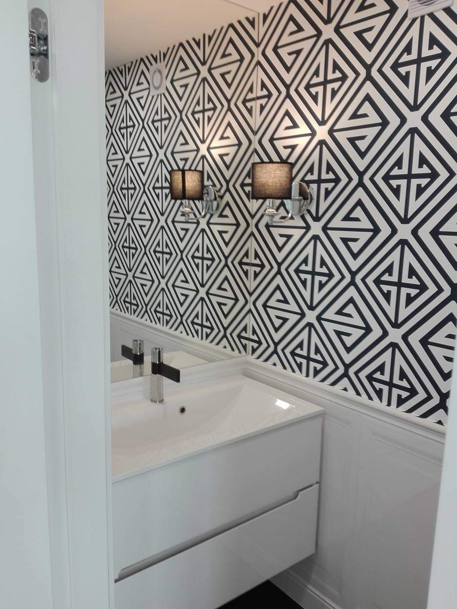 Nowoczesna łazienka z białą szafką pod umywalką, baterią w kolorze chromu i lustrem odbijającym wzorzystą tapetę w geometryczne czarno-białe motywy, oświetloną przez dwa kinkiety z abażurami...