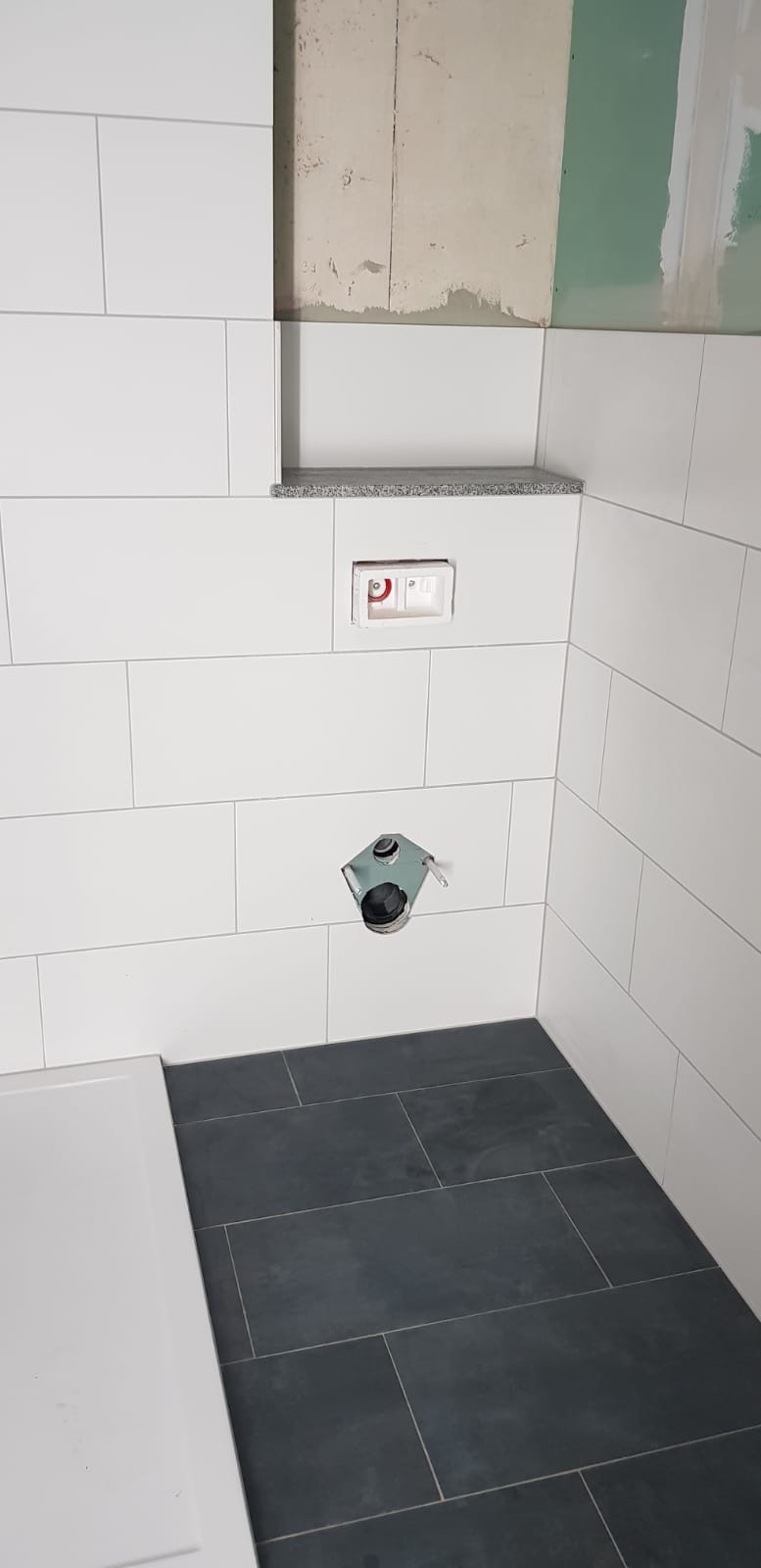 Fragment łazienki w trakcie prac wykończeniowych: białe płytki ścienne, grafitowa podłoga z płytek, zamontowany stelaż podtynkowy WC, widoczne niedokończone fragmenty ściany.