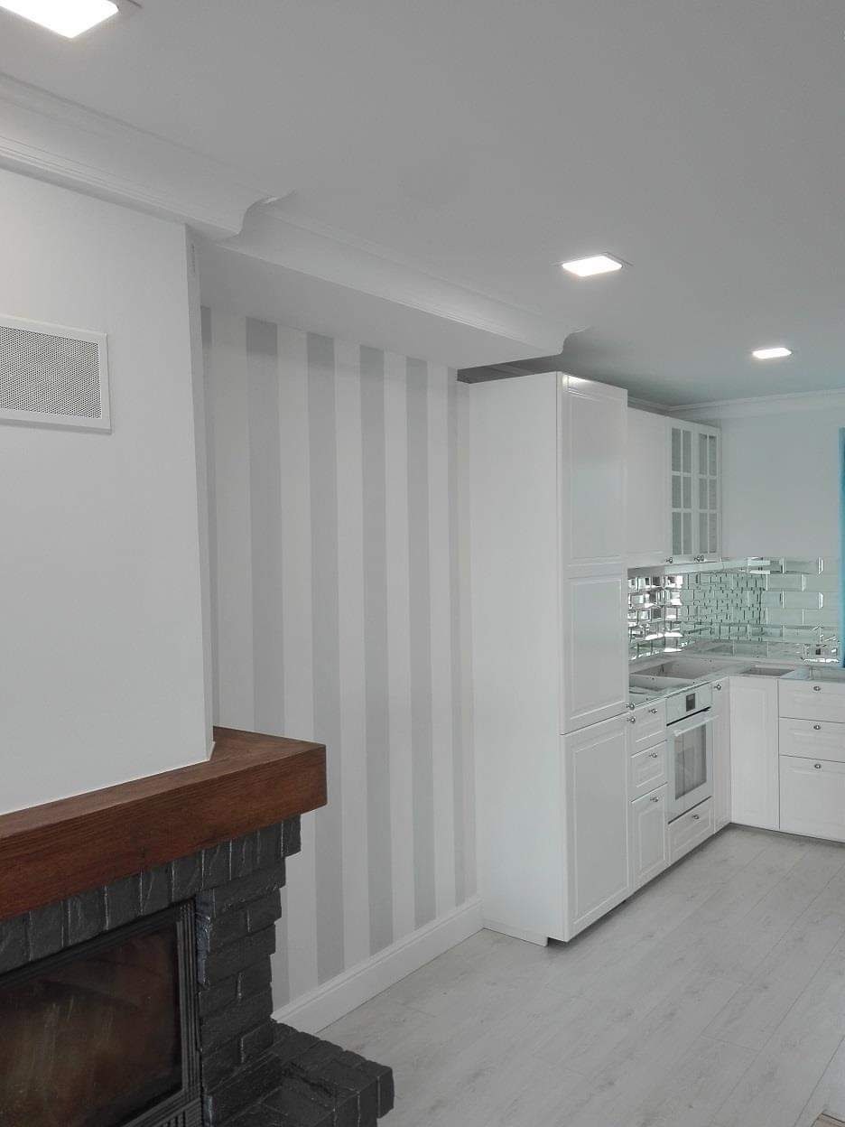 Wnętrze salonu z kominkiem i aneksem kuchennym: ściana w szare pasy, białe meble kuchenne z lustrzanymi płytkami nad blatem, drewniana obudowa kominka.