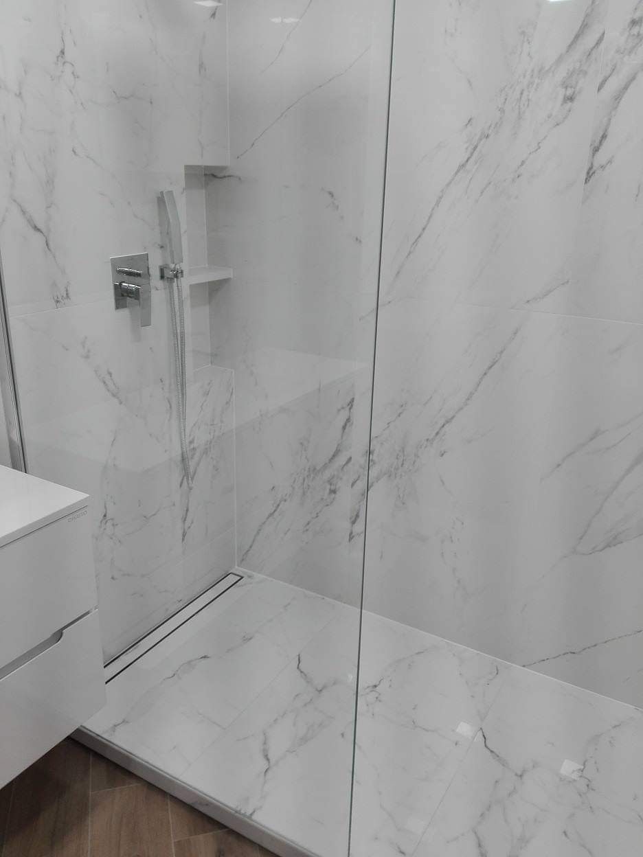 Nowoczesna kabina prysznicowa walk-in z białymi płytkami imitującymi marmur, chromowaną armaturą i odpływem liniowym.