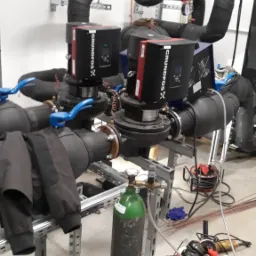 Dwa czarne pompy Grundfos X, połączone rurami z niebieskimi zaworami, zamontowane na metalowej konstrukcji. Obok butla z gazem spawalniczym i skrzynka z narzędziami.