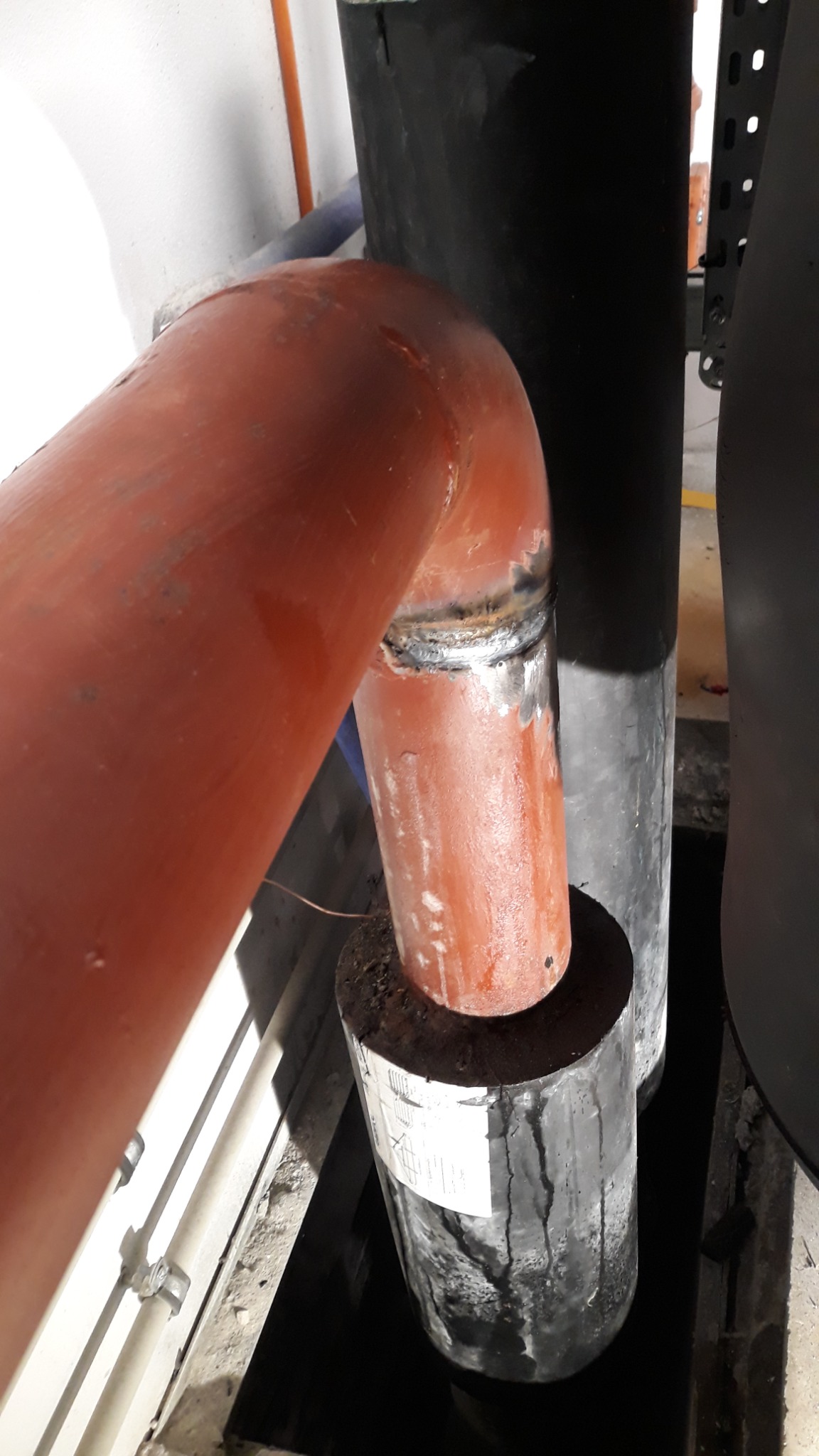 Pomarańczowy spawany rurociąg z widocznymi śladami spawania, wsparty na czarnym, cylindrycznym elemencie w przestrzeni technicznej.