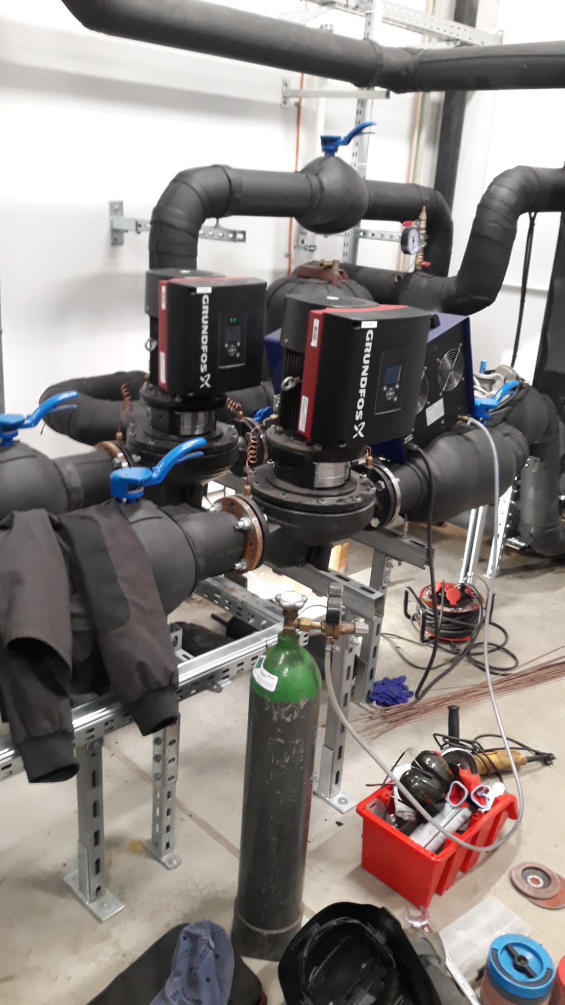 Dwa czarne pompy Grundfos X, połączone rurami z niebieskimi zaworami, zamontowane na metalowej konstrukcji. Obok butla z gazem spawalniczym i skrzynka z narzędziami.