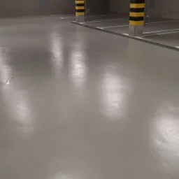 Wypolerowana, jasnoszara posadzka w garażu podziemnym z betonowymi słupami oznaczonymi czarno-ż&oacute;łtymi pasami, widoczne linie wyznaczające miejsca parkingowe i oświetlenie sufitowe.