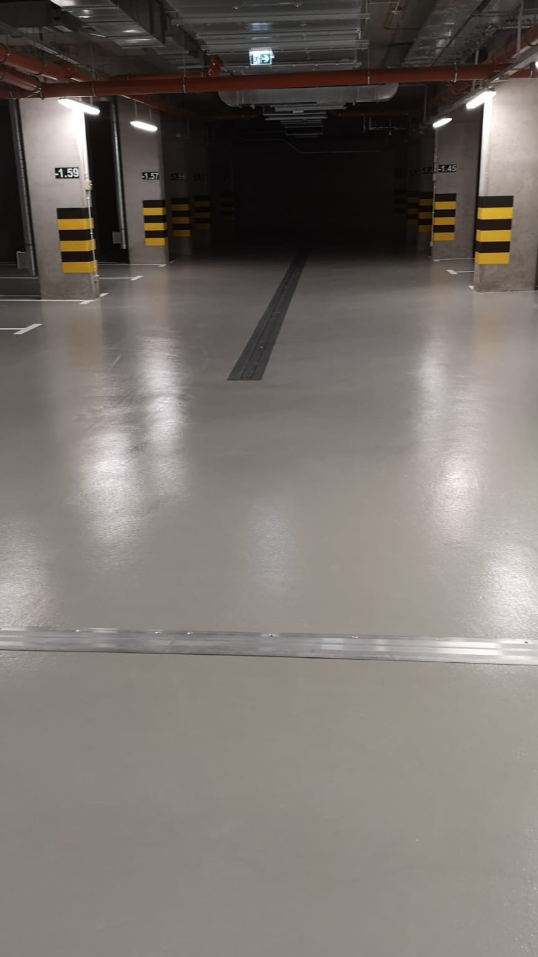 Błyszcząca, szara posadzka w garażu podziemnym, z widocznymi słupami oznaczonymi numerami i żółto-czarnymi pasami ostrzegawczymi oraz centralnie umieszczonym odpływem liniowym, oświetlona przez...