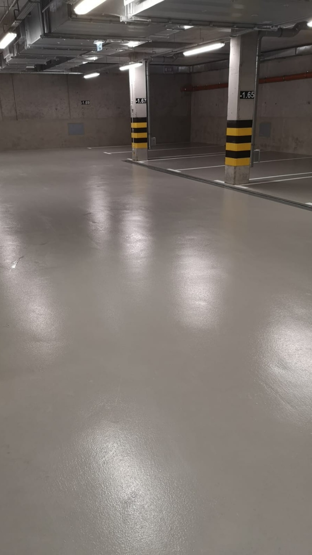 Wypolerowana, jasnoszara posadzka w garażu podziemnym z betonowymi słupami oznaczonymi czarno-żółtymi pasami, widoczne linie wyznaczające miejsca parkingowe i oświetlenie sufitowe.