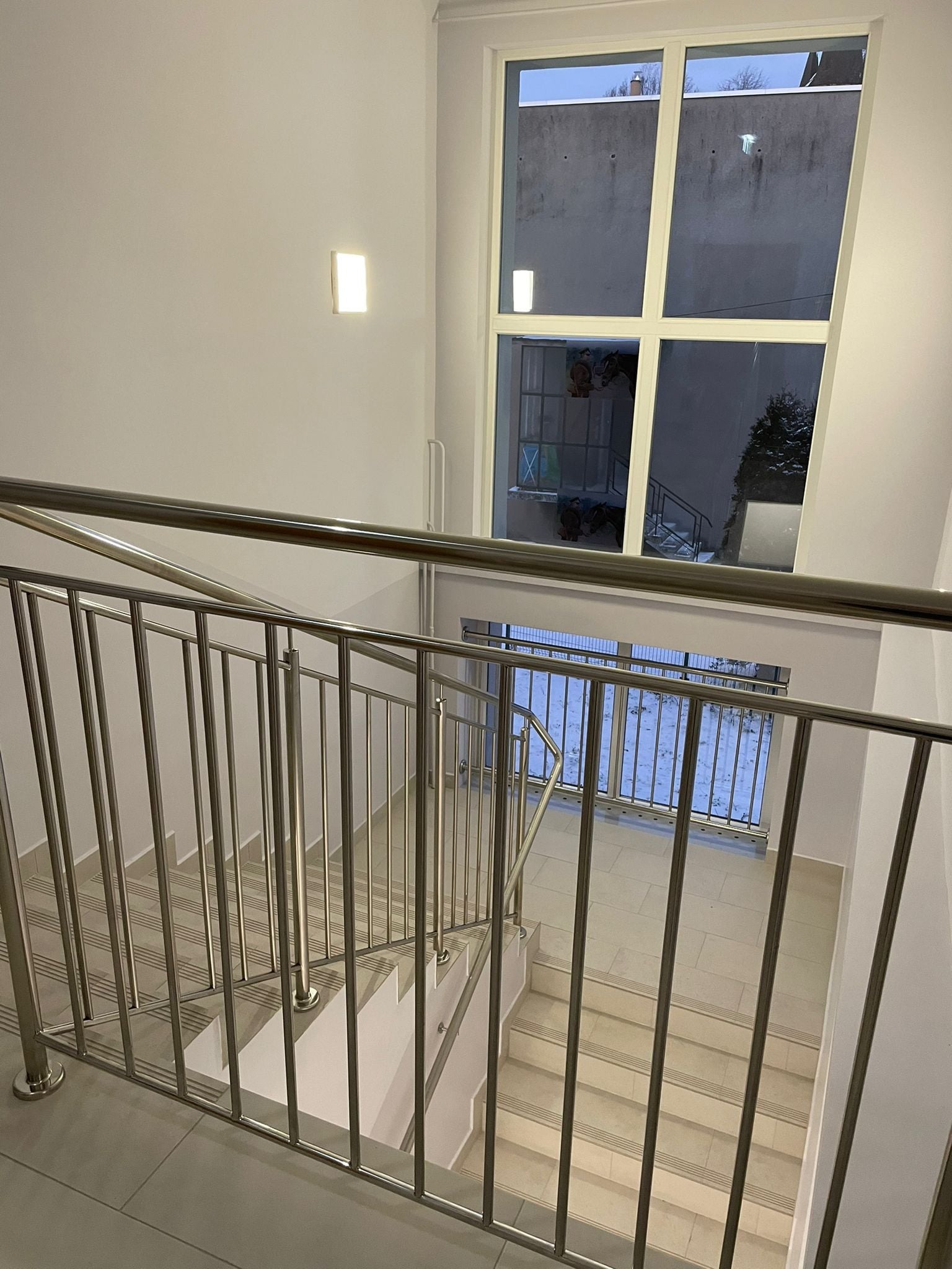 Klatka schodowa z metalową balustradą widziana z góry, jasne płytki na schodach, okno z widokiem na ośnieżony ogród i budynek w tle.