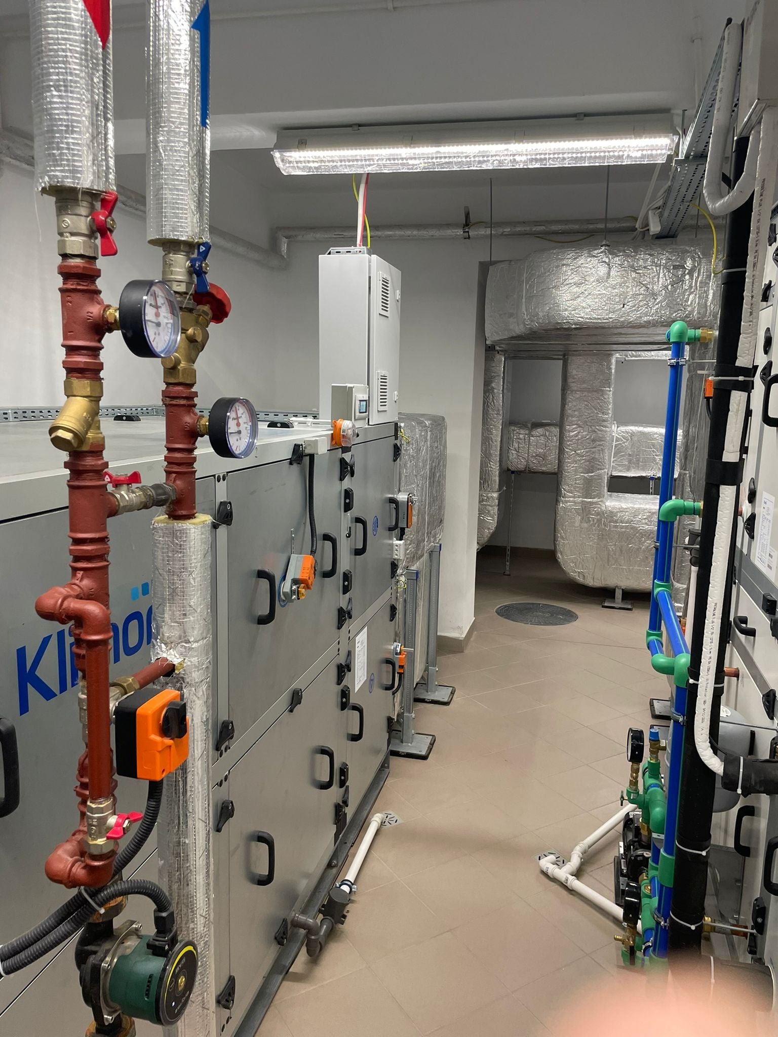 Wnętrze techniczne z systemem wentylacji i rurami w kolorach czerwonym, niebieskim i zielonym, widoczne manometry i zawory, metalowe kanały wentylacyjne w izolacji.