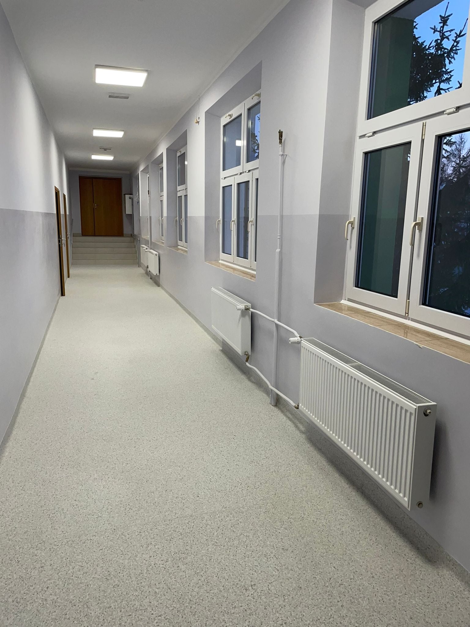 Długi korytarz w budynku użyteczności publicznej z szarą podłogą lastryko, białymi ścianami z szarym pasem, białymi grzejnikami pod oknami i drewnianymi drzwiami w oddali.