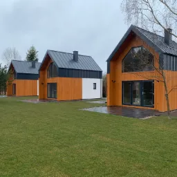 Trzy nowoczesne domy szkieletowe z drewnianą elewacją i grafitowym dachem na tle zielonego trawnika i pochmurnego nieba.