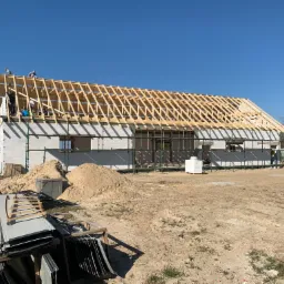 Wznoszenie budynku z białych bloczków, z widoczną drewnianą konstrukcją dachu i pracownikami na górze, materiały budowlane składowane w pobliżu.