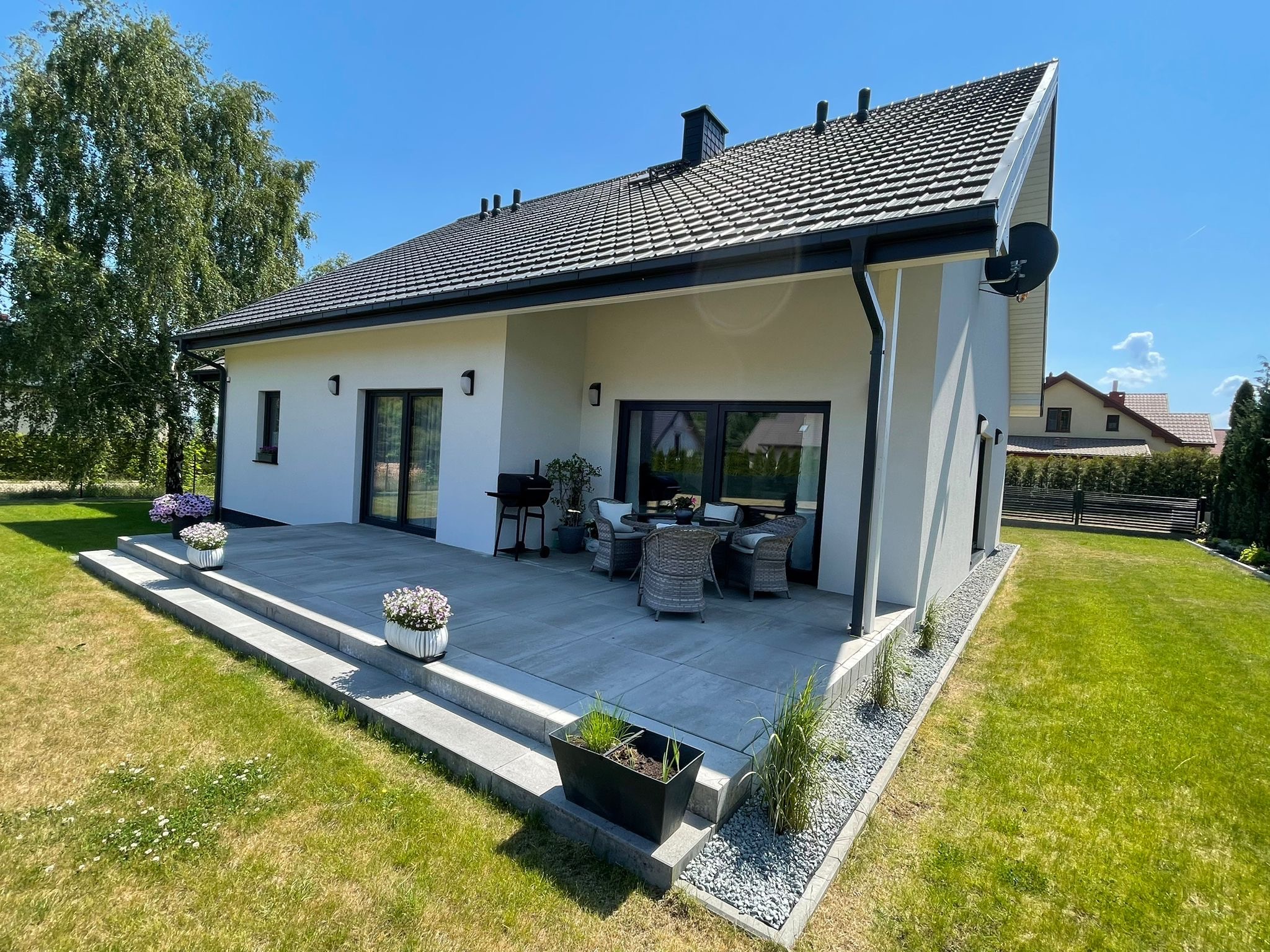 Nowoczesny dom jednorodzinny z ciemną dachówką karpiówką, jasną elewacją i tarasem z meblami ogrodowymi, otoczony zielonym trawnikiem.