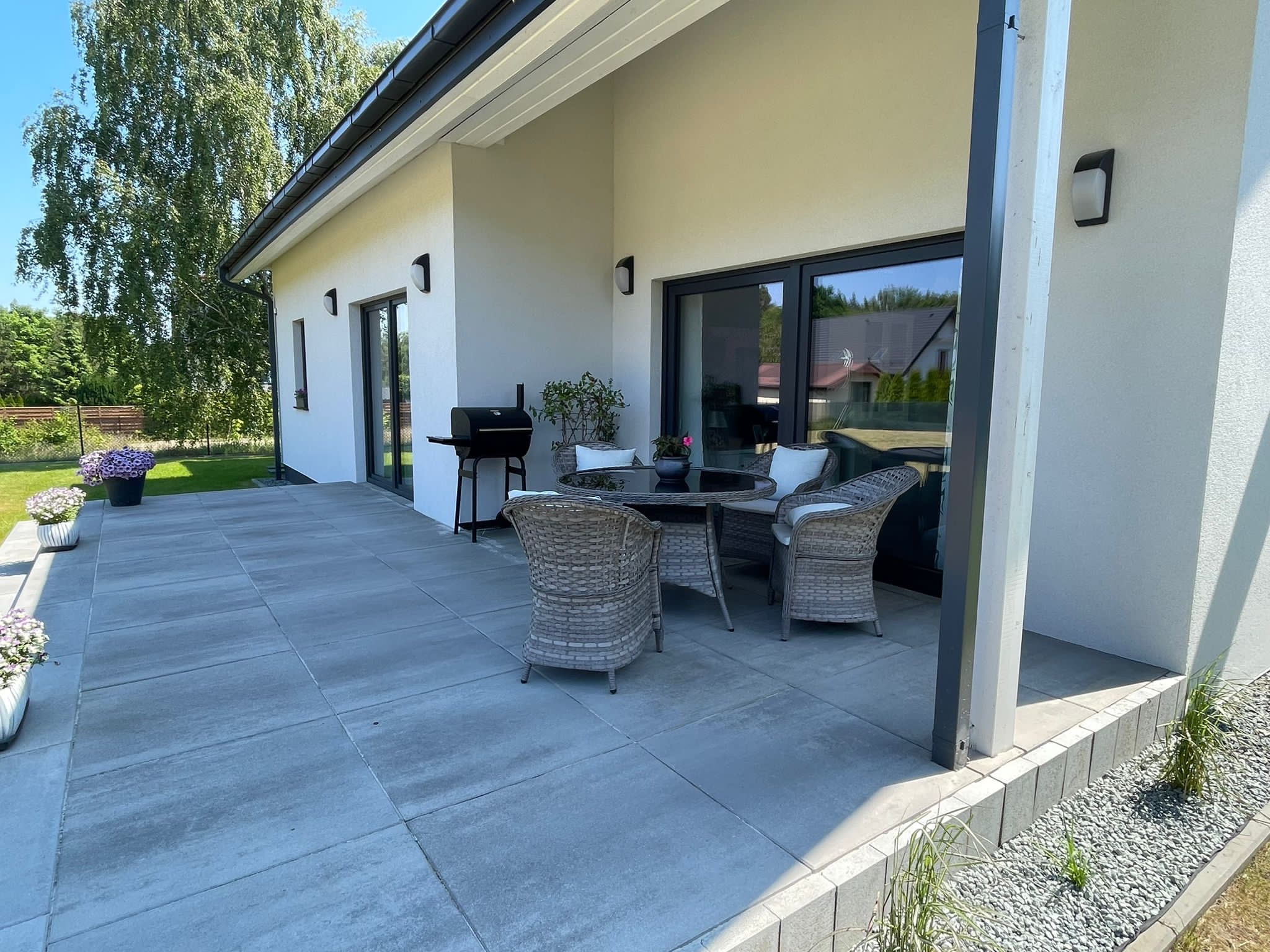 Nowoczesny taras z szarych płyt betonowych, wyposażony w rattanowe meble ogrodowe, grill i kwiaty w donicach, przylegający do białego domu z ciemnymi oknami.