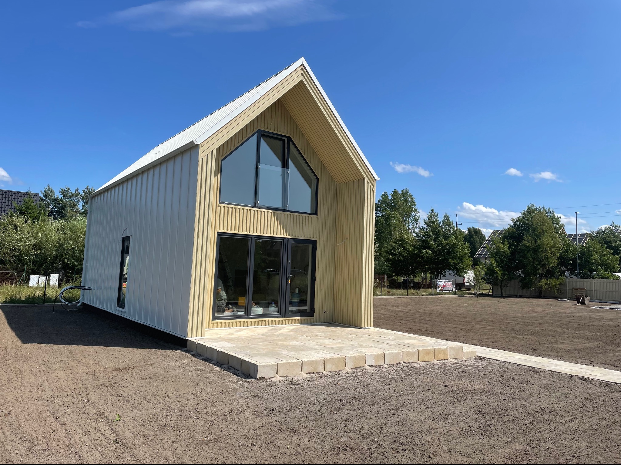 Nowoczesny, parterowy budynek z jasną elewacją z pionowych desek, dużymi oknami i płaskim dachem, otoczony przygotowanym terenem pod trawnik.