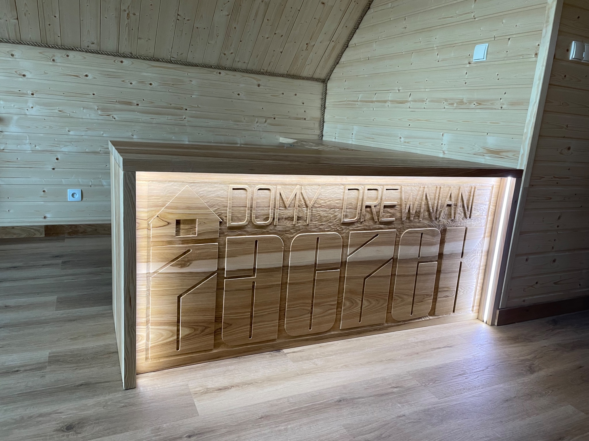 Wnętrze domu szkieletowego z wykończeniem drewnianym, widoczny fragment ściany z podświetlanym napisem 'DOMY DREWNIANE 2024'.