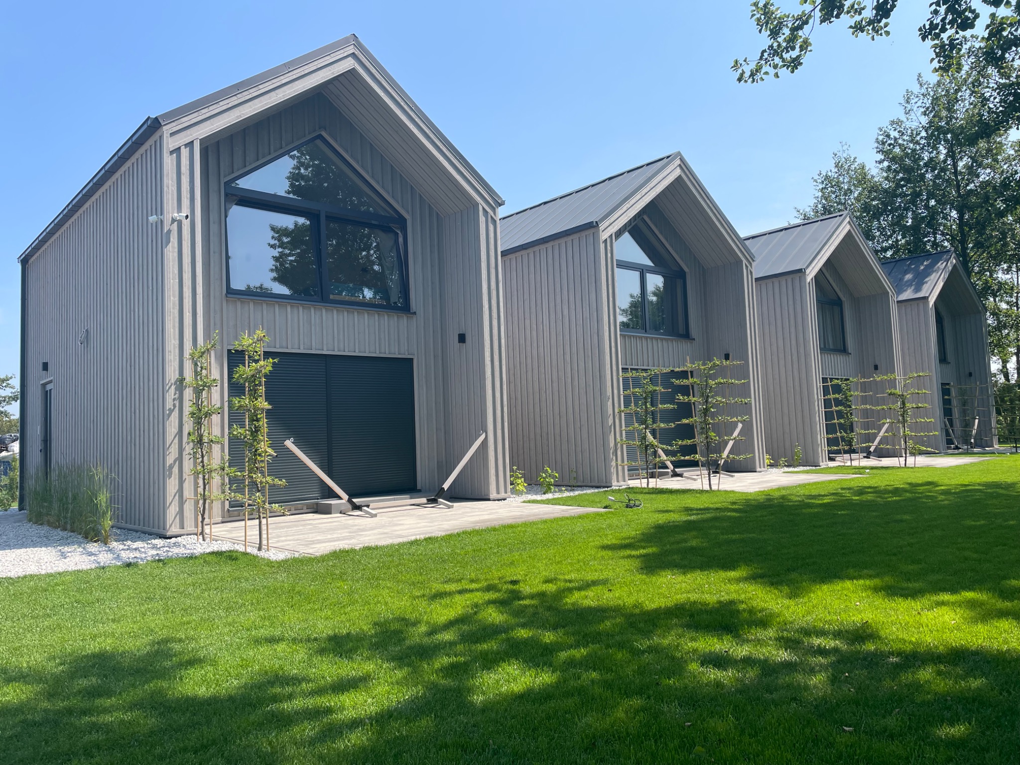 Rząd nowoczesnych domów szkieletowych z szarym metalowym dachem i dużymi trójkątnymi oknami, otoczonych zielonym trawnikiem i młodymi drzewkami.