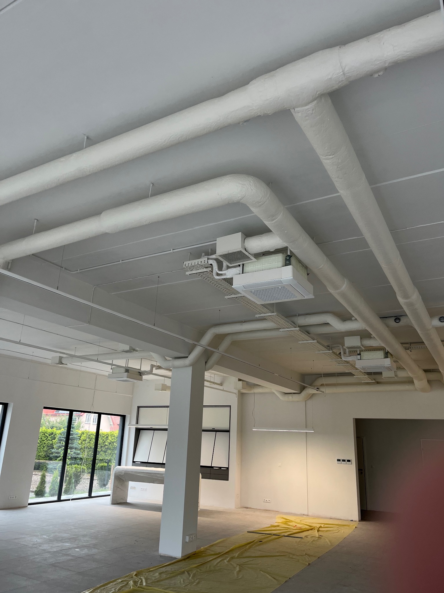 Białe rury wentylacyjne i klimatyzatory kasetonowe pod sufitem w jasnym, przestronnym wnętrzu biurowym z dużymi oknami i żółtą folią ochronną na podłodze.