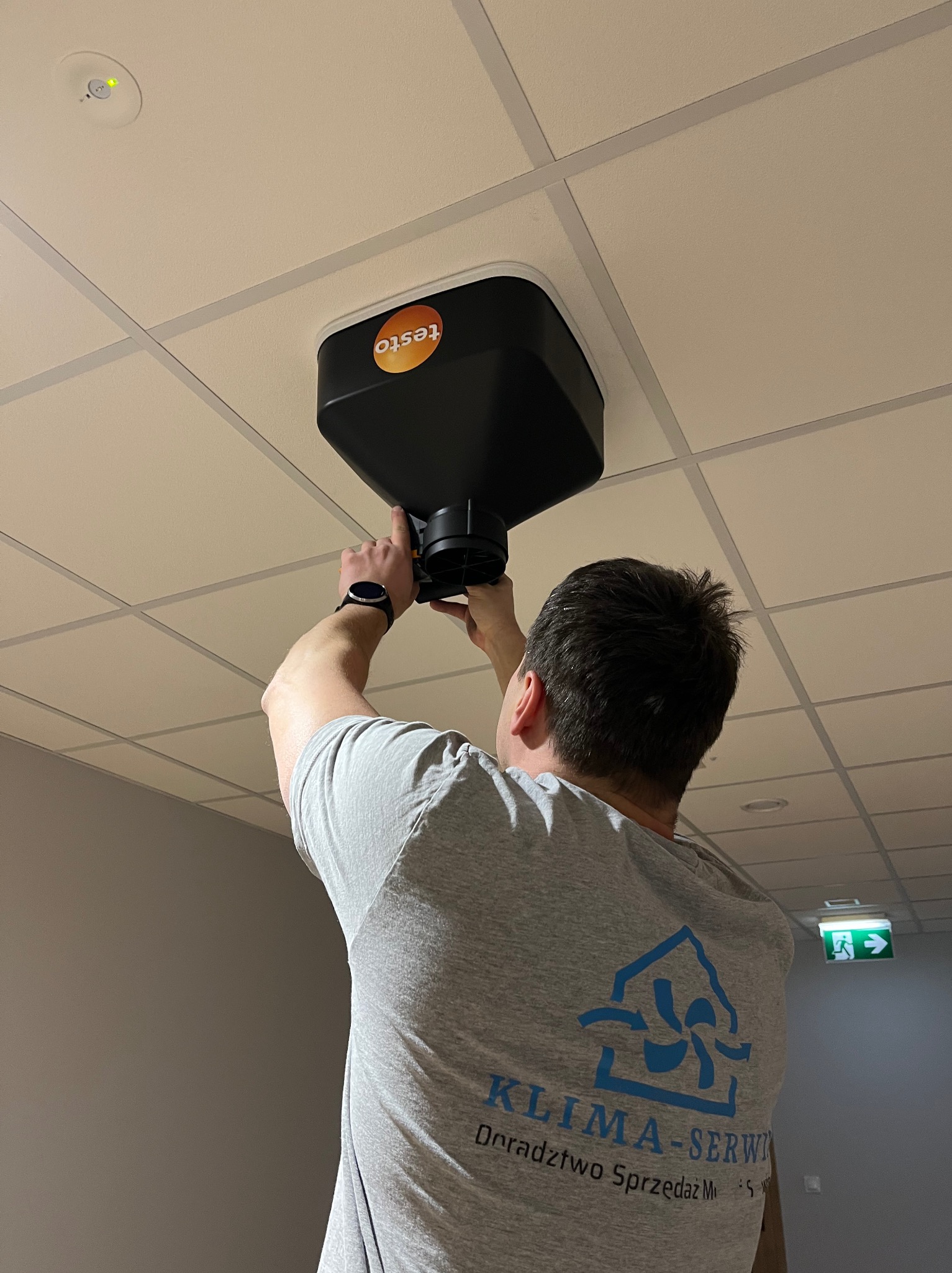 Instalator z logiem firmy na koszulce montuje anemostat Testo na suficie kasetonowym w biurze. Widoczny czujnik dymu i znak ewakuacyjny.