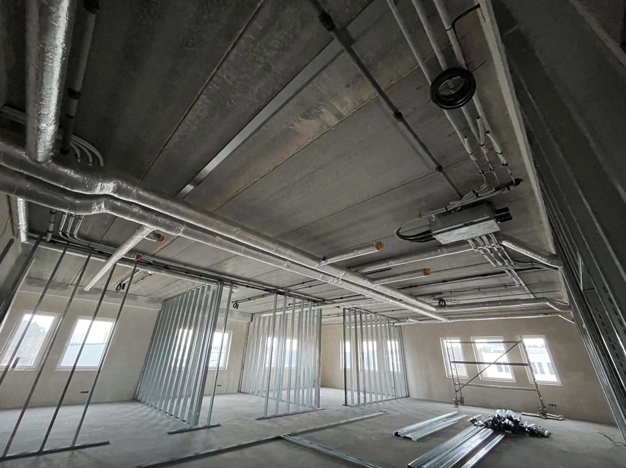 Wnętrze w stanie surowym z widoczną instalacją wentylacyjną pod sufitem i konstrukcją ścian działowych z profili stalowych. Widoczne okna i surowy betonowy sufit.
