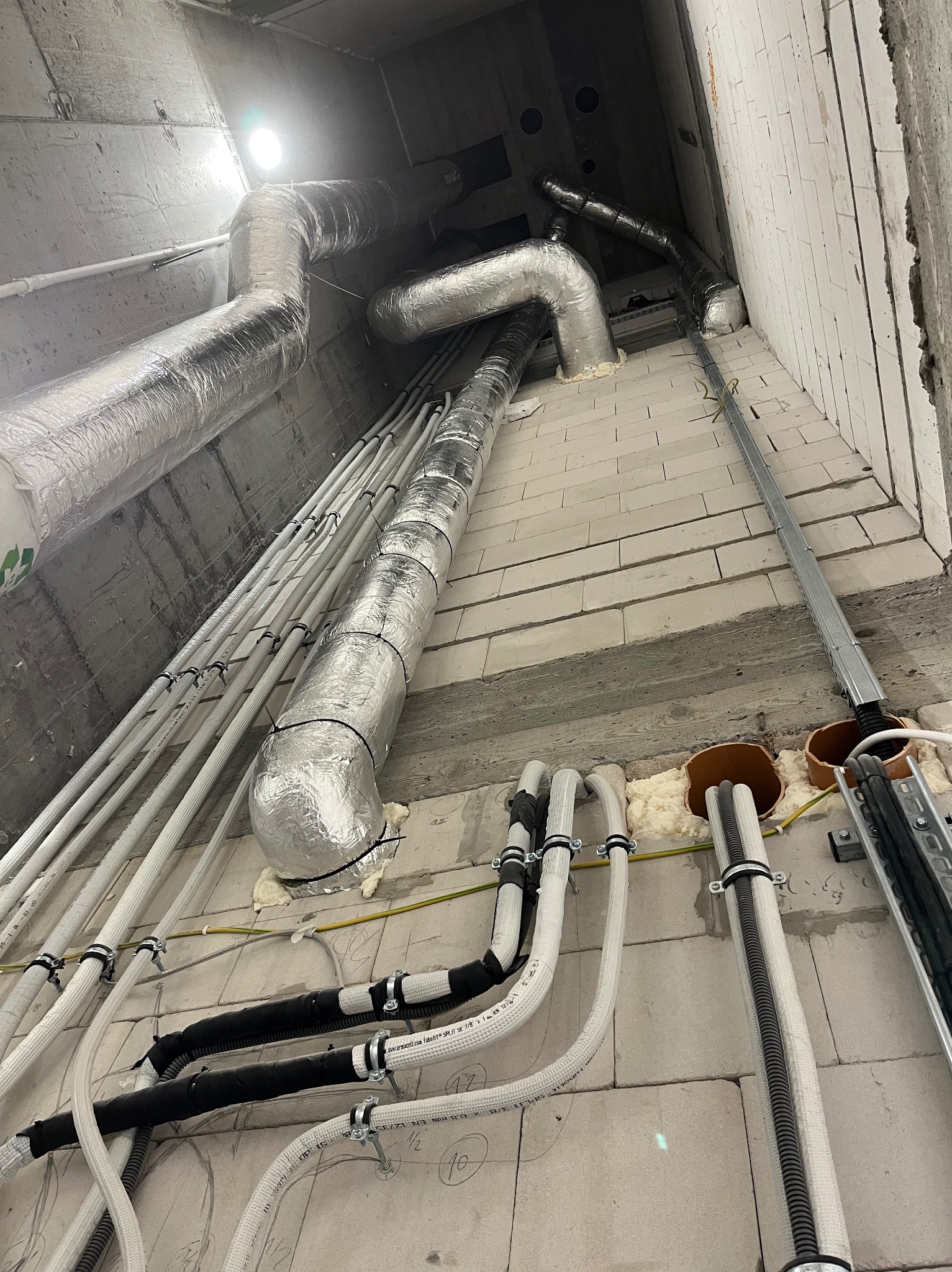 Montaż instalacji wentylacyjnej z rurami w izolacji aluminiowej i przewodami elektrycznymi na surowej ścianie z betonu komórkowego w budynku.