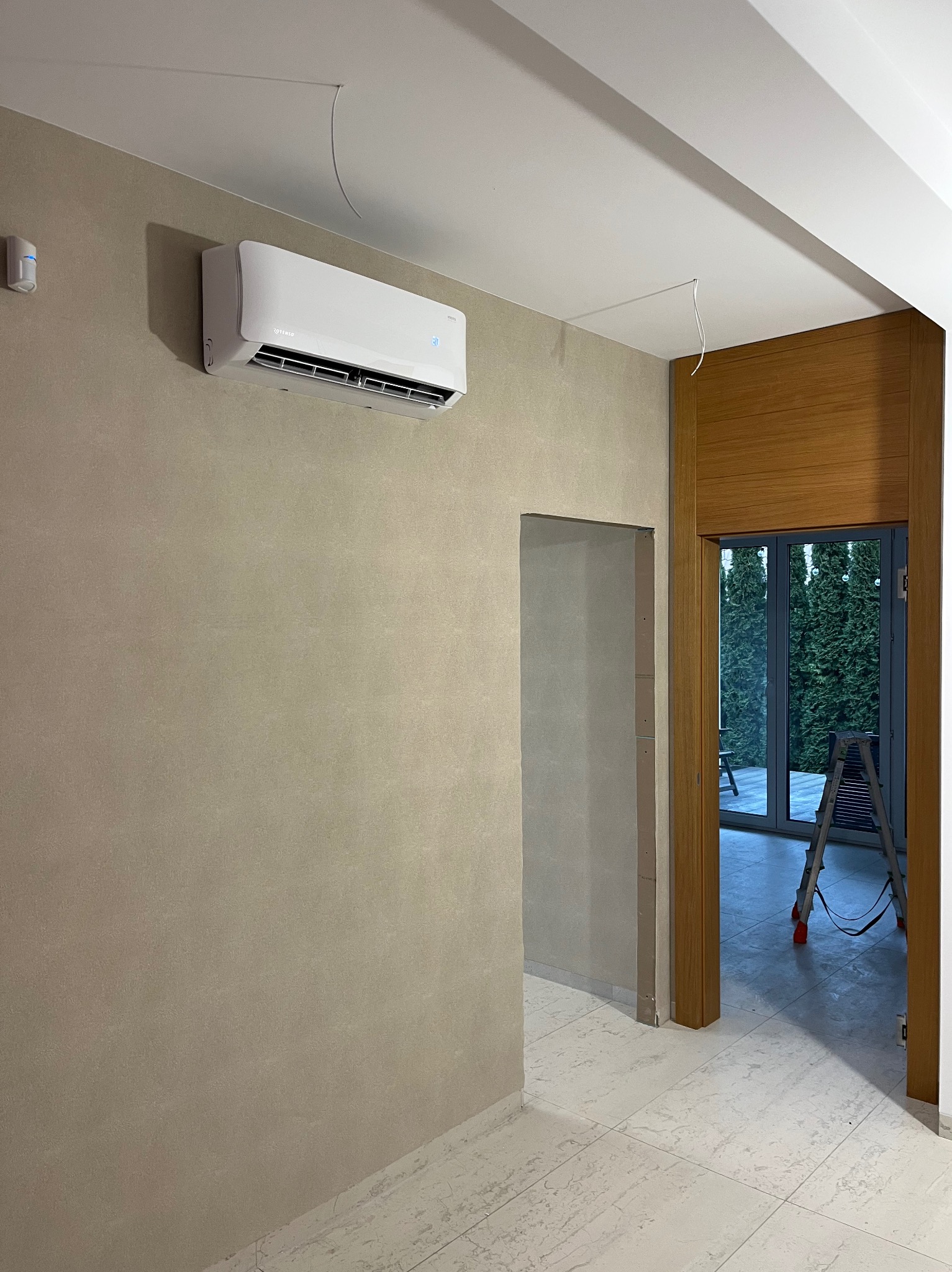 Biały klimatyzator Rotenso zamontowany na beżowej ścianie w jasnym wnętrzu. Widoczne przewody i drabina w tle sugerują etap instalacji.