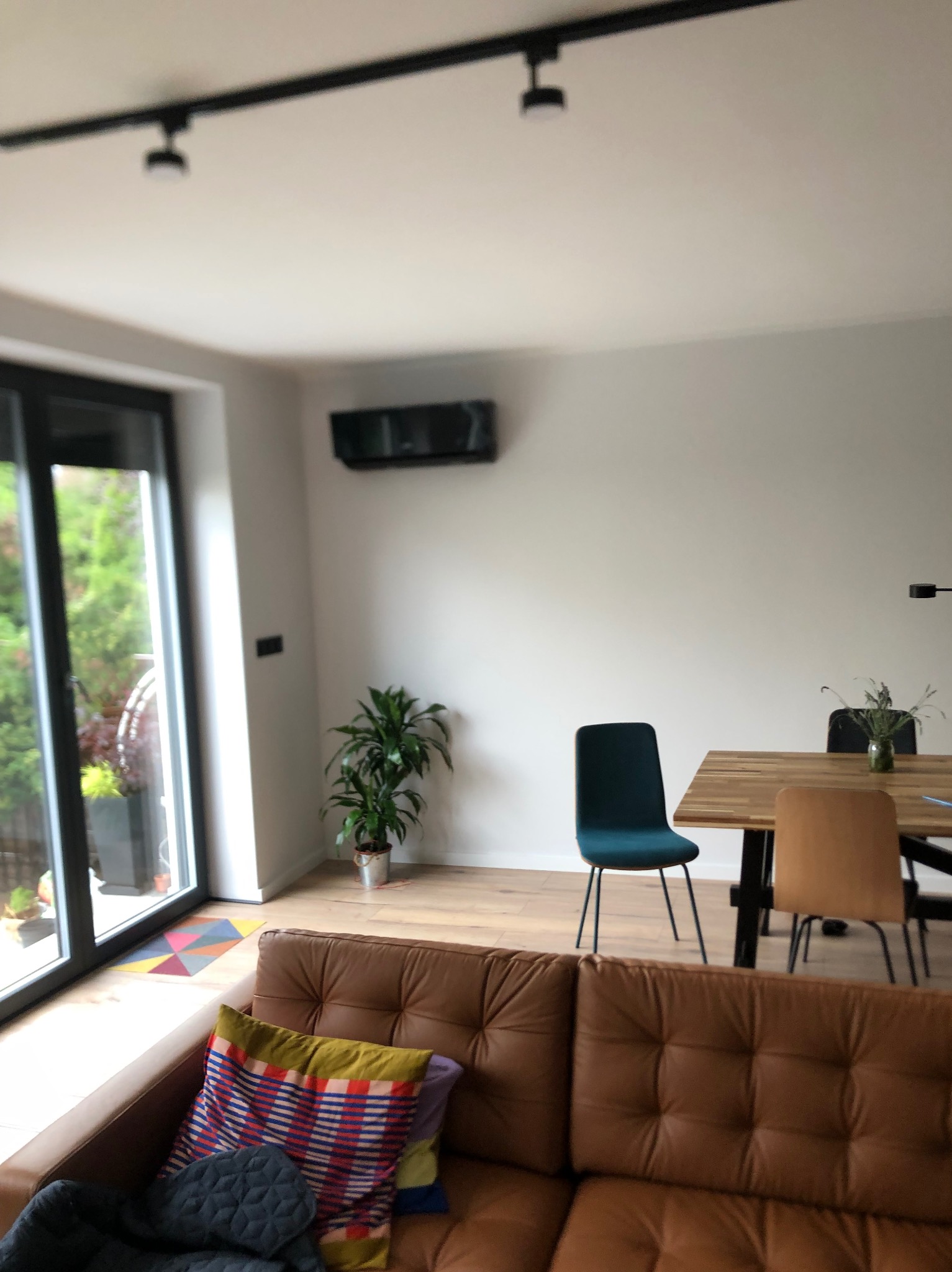 Nowoczesny salon z brązową sofą, drewnianym stołem i klimatyzatorem ściennym. Minimalistyczny design, jasne wnętrze z roślinami doniczkowymi i dużym oknem.