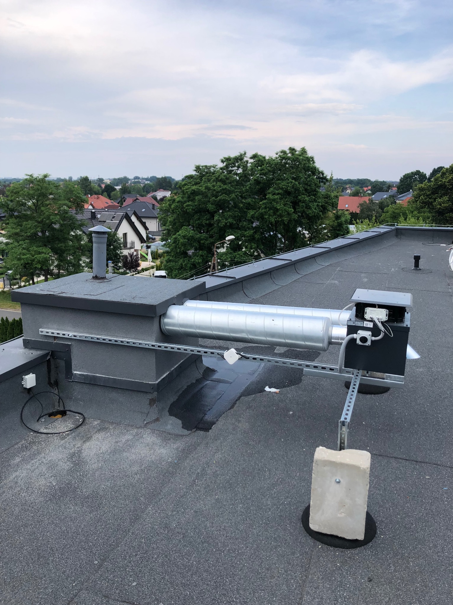 Instalacja wentylacyjna na dachu budynku z widokiem na okoliczne domy i zieleń. Elementy montażowe i rury wentylacyjne w kolorze srebrnym i szarym.