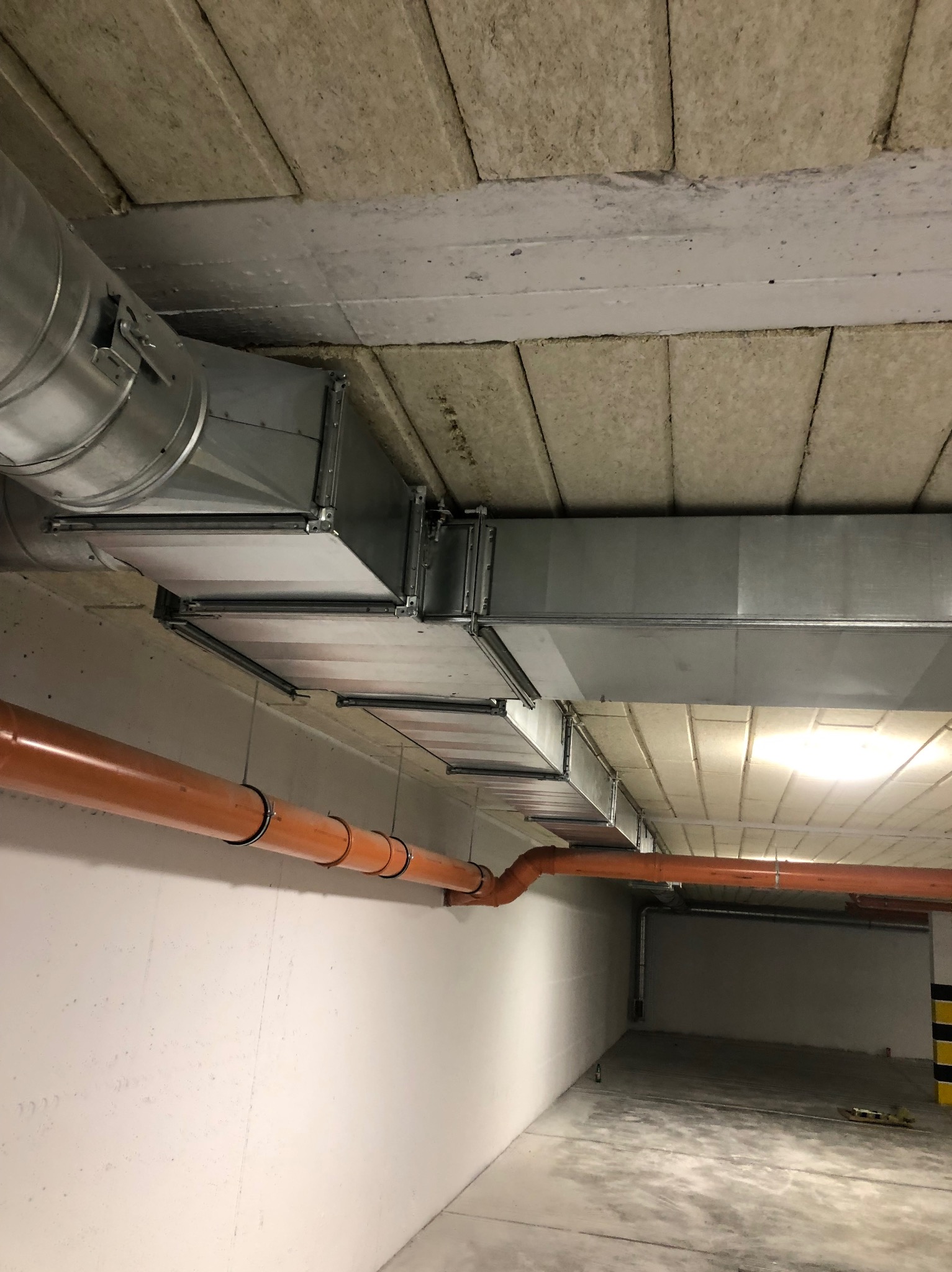 Instalacja wentylacyjna z metalowych kanałów i pomarańczowej rury w surowym wnętrzu, widok z dołu, oświetlenie sufitowe.