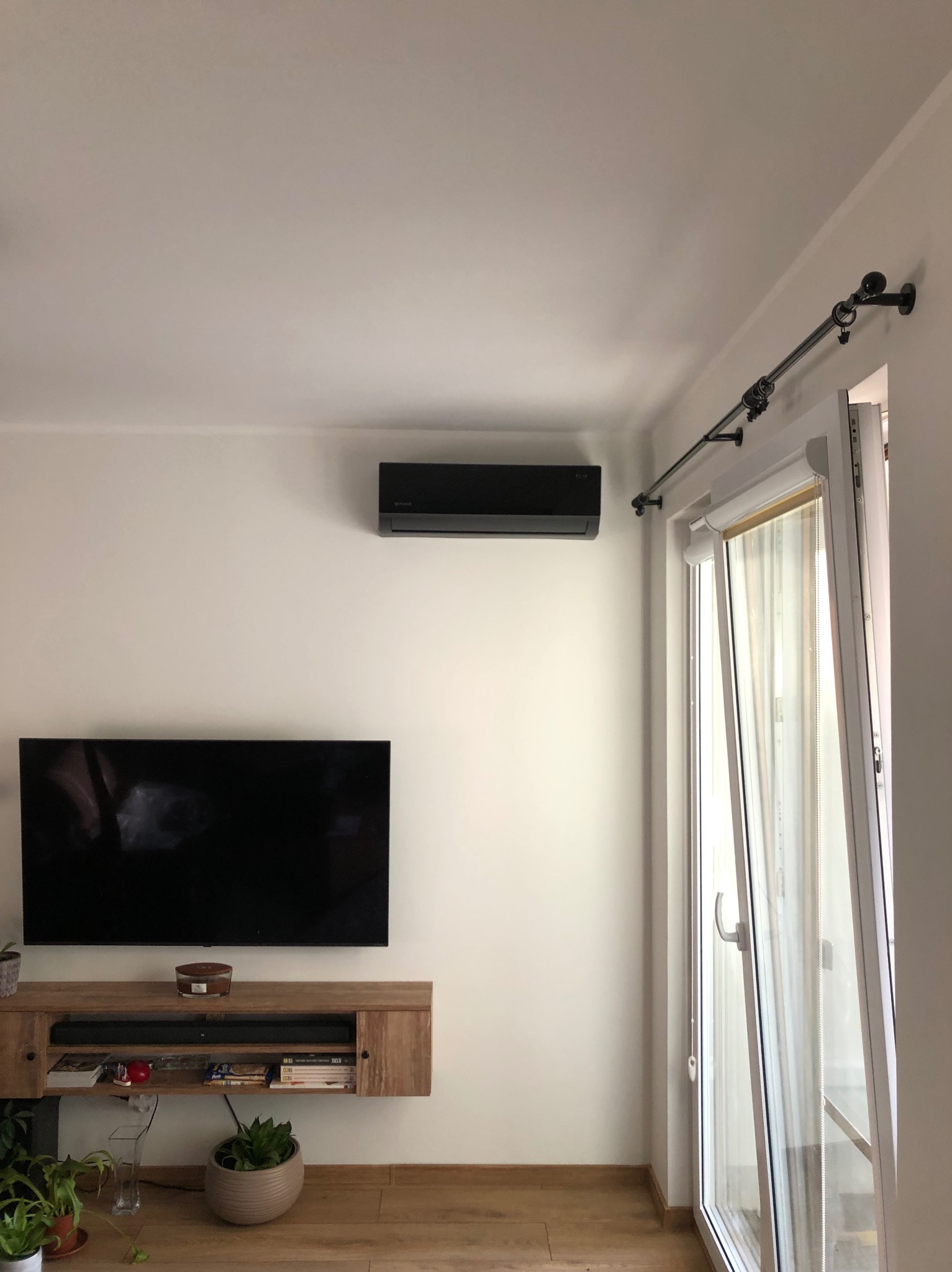 Czarny klimatyzator ścienny w nowoczesnym salonie w Toruniu. Widoczny telewizor i balkon. Minimalistyczny design, jasne ściany, drewniane elementy.