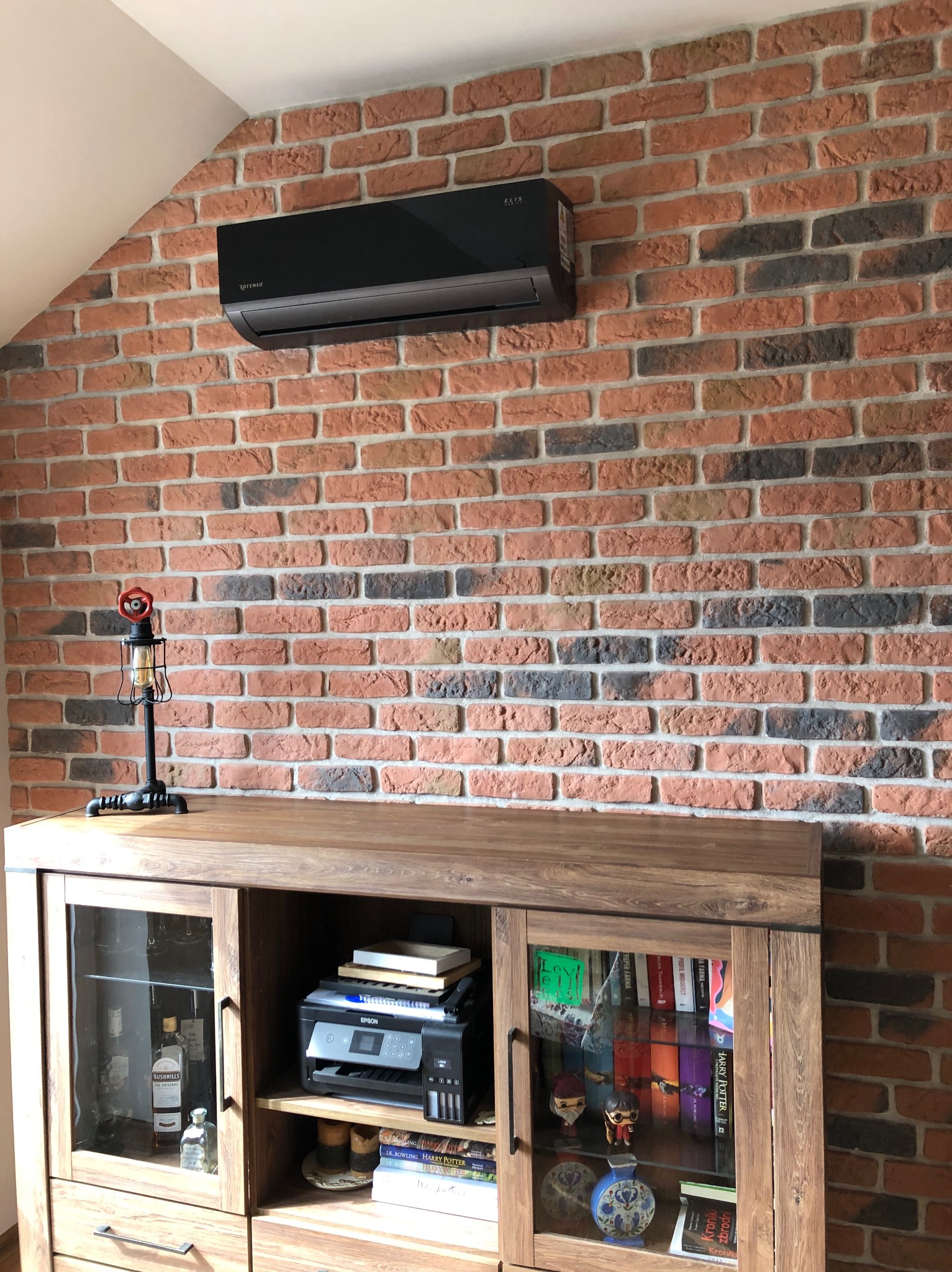 Czarny klimatyzator Rotenso Elis zamontowany na ścianie z cegły w pomieszczeniu z drewnianą komodą. Widoczne książki i dekoracje.