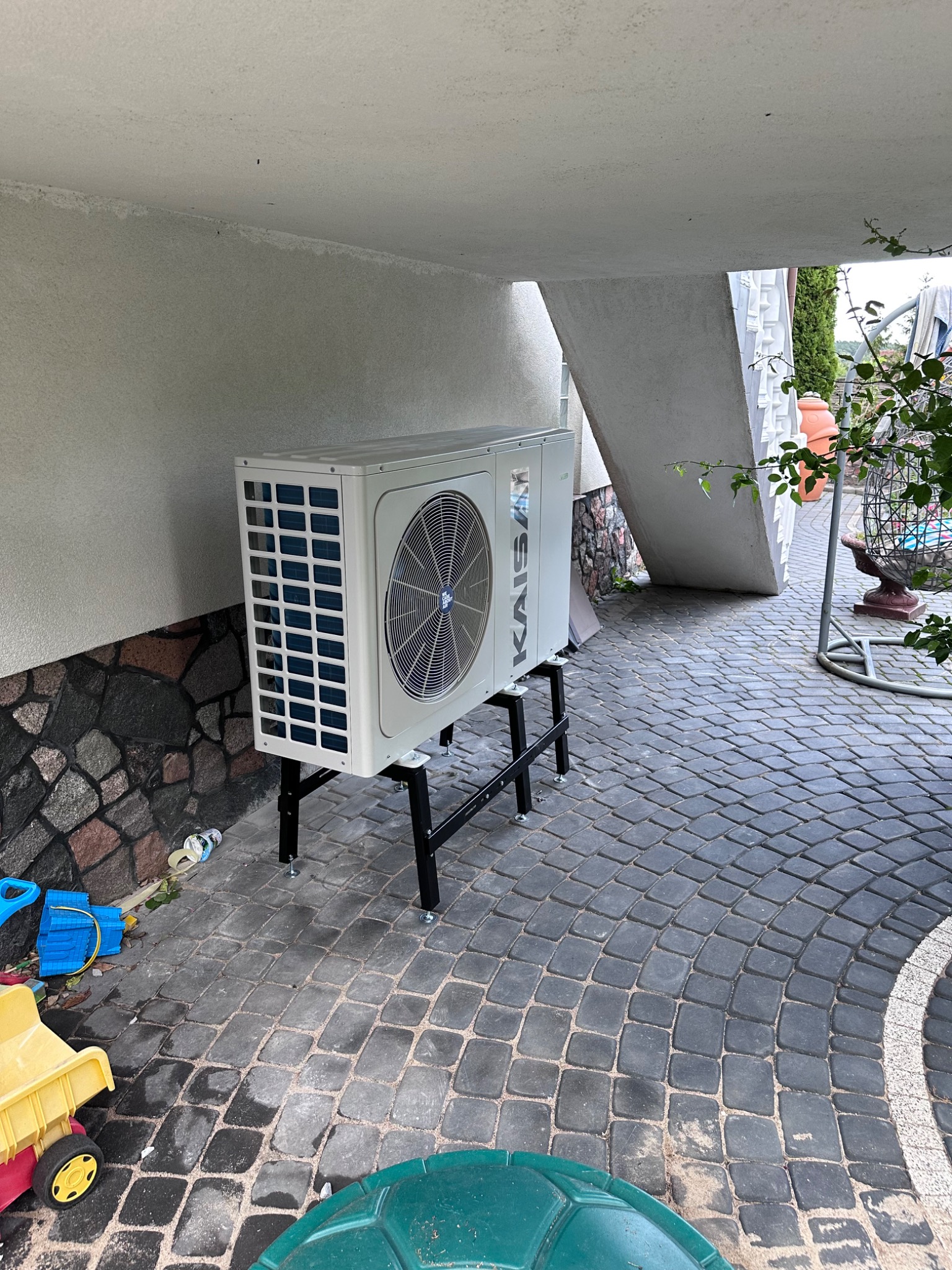 Biała pompa ciepła marki KAISAI zamontowana na czarnym, metalowym stelażu na kostce brukowej, obok ściany z kamienia i dziecięcych zabawek.