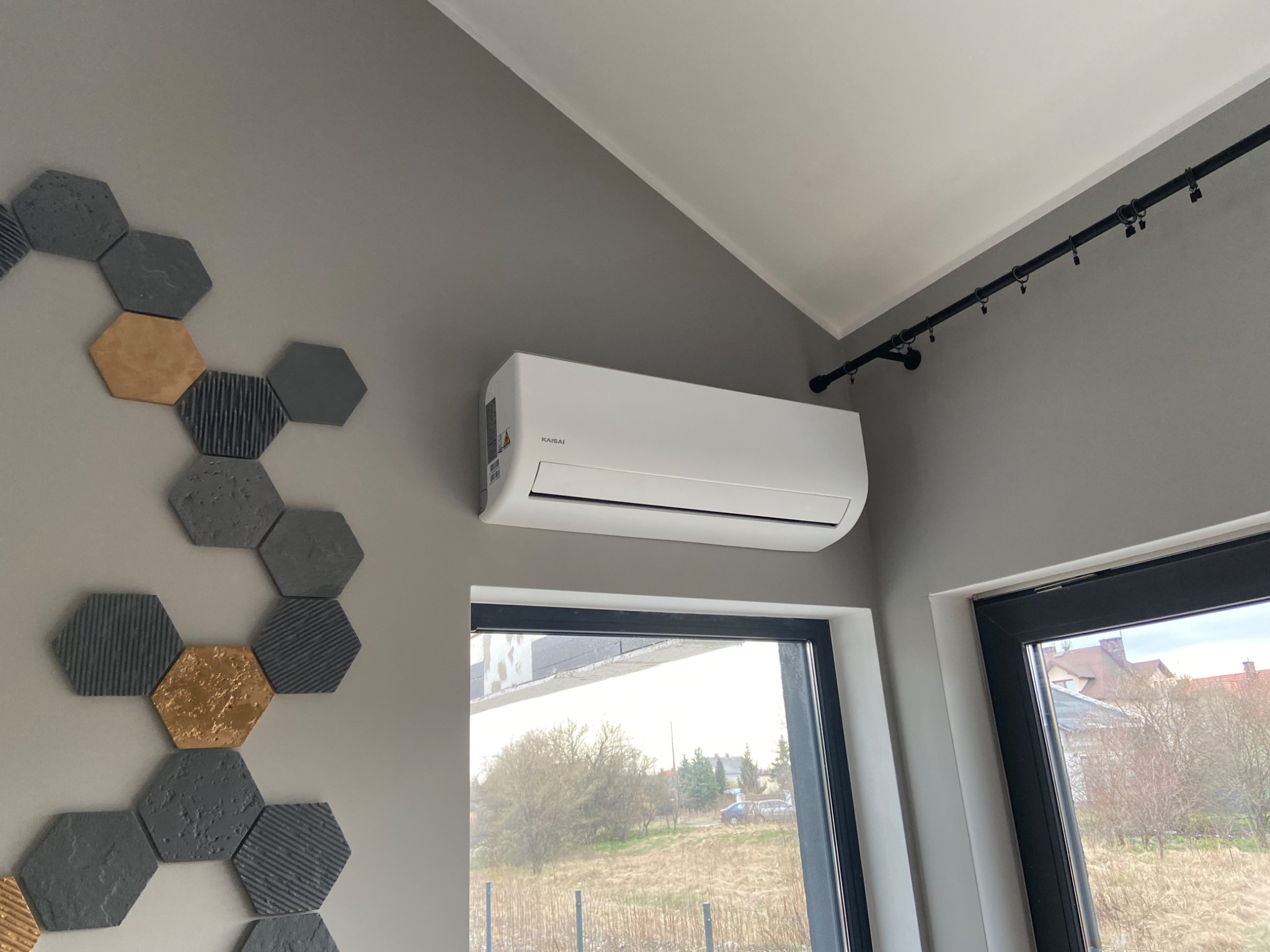 Biała jednostka klimatyzacyjna Kaisai zamontowana na szarej ścianie nad oknem, z dekoracyjnymi heksagonami w odcieniach szarości i złota po lewej stronie oraz czarną szyną oświetleniową pod sufitem.