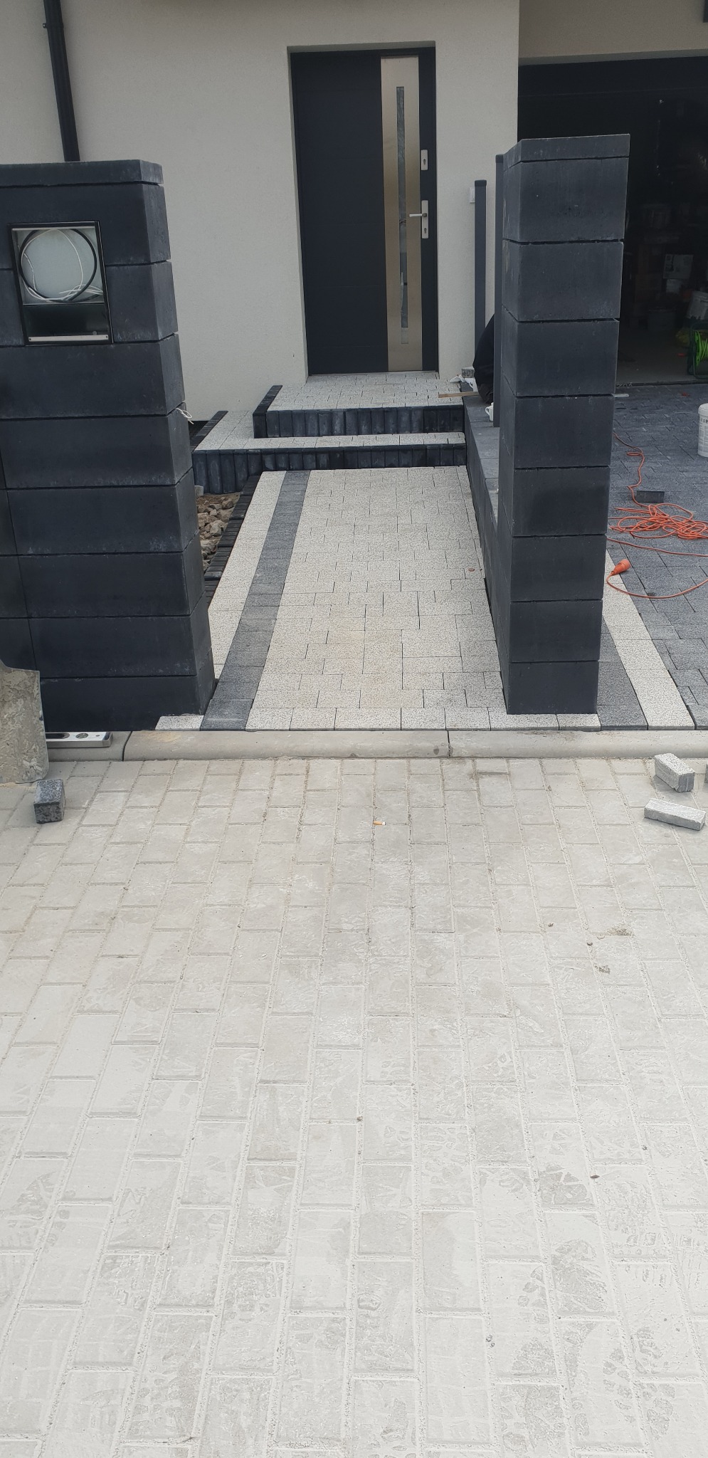 Nowo ułożona, jasnoszara kostka brukowa przed domem z ciemnymi elementami obrzeży i schodów, widoczne słupki ogrodzeniowe z ciemnej kostki i fragment zwiniętego kabla elektrycznego.