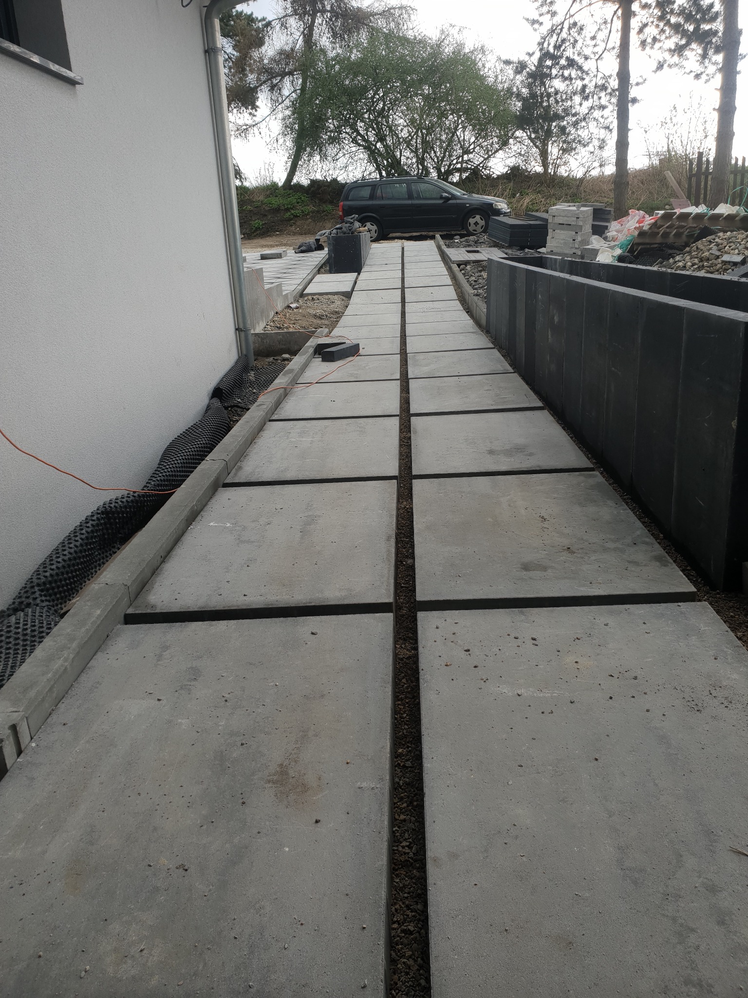 Ułożone betonowe płyty chodnikowe tworzą ścieżkę wzdłuż ściany budynku, widoczna czarna folia kubełkowa przy fundamencie oraz zaparkowany samochód w tle.