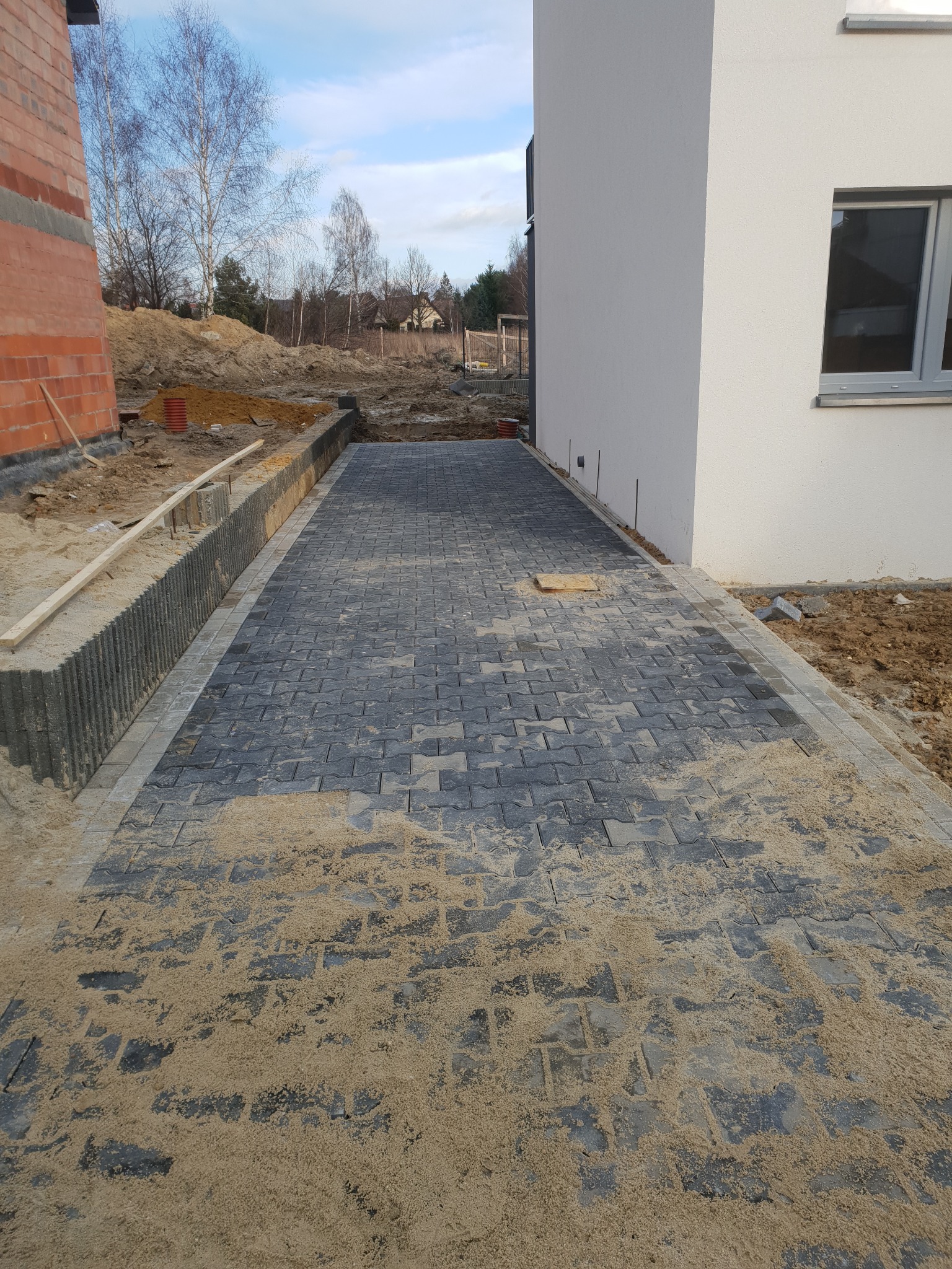 Świeżo ułożona, szara kostka brukowa z piaskiem w Zabrzu, pomiędzy budynkiem a ogrodzeniem, w trakcie prac wykończeniowych.