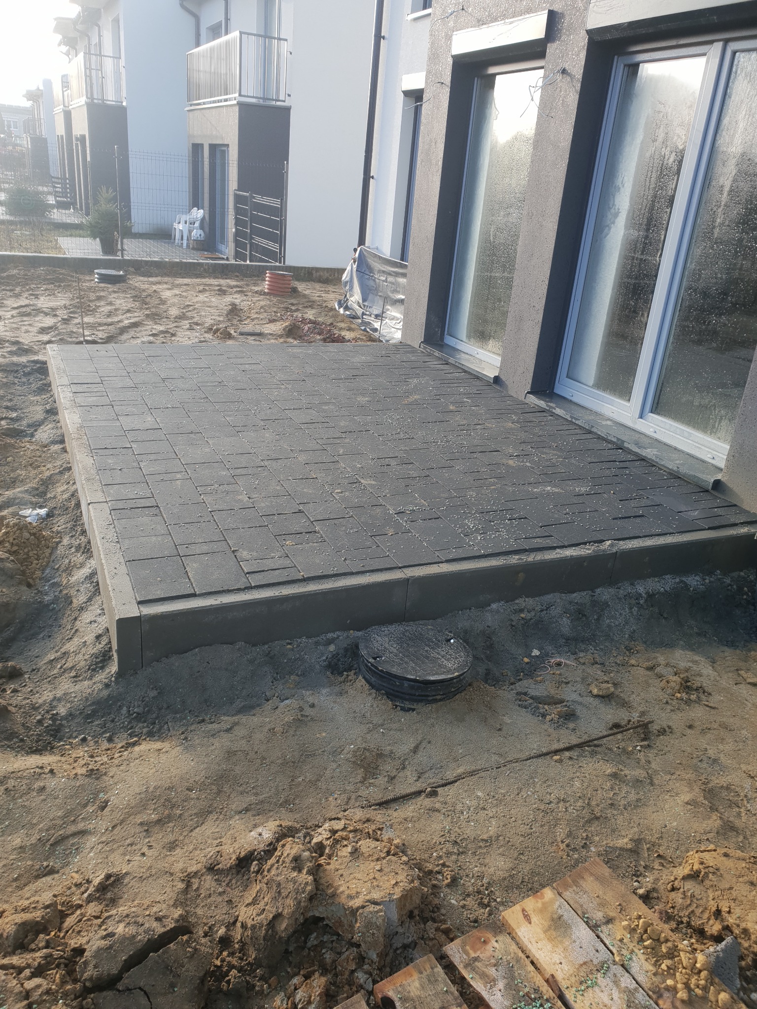 Nowo ułożony, prostokątny taras z ciemnoszarej kostki brukowej przylegający do ściany budynku z oknami, otoczony piaskiem i ziemią, z widoczną studzienką kanalizacyjną i fragmentem palety.