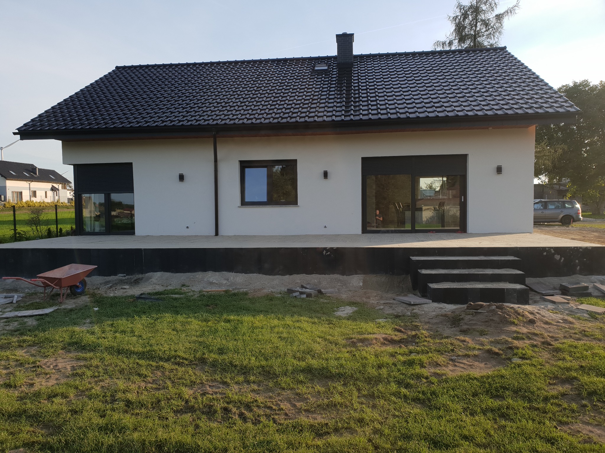 Nowoczesny dom z tarasem wyłożonym szarą kostką brukową, widoczne schody wejściowe i taczka na trawniku, teren w trakcie prac wykończeniowych.