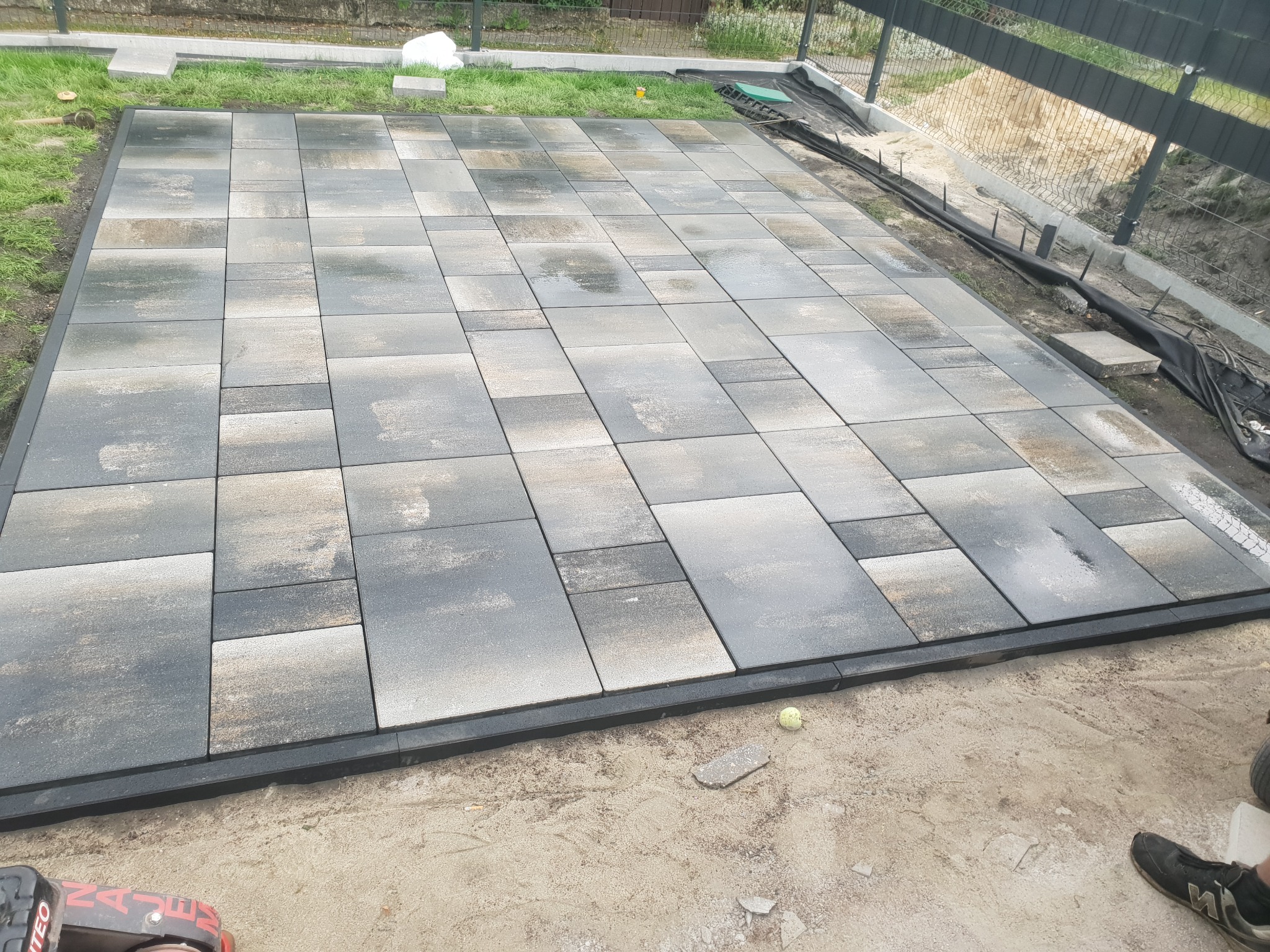 Świeżo ułożona, wilgotna kostka brukowa w odcieniach szarości, z czarnym obrzeżem, na podsypce piaskowej, widoczny fragment buta i zagęszczarka w lewym dolnym rogu, w tle ogrodzenie z siatki.