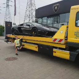 Czarny Lamborghini Aventador na żółtej lawecie pomocy drogowej, obok stoi chłopiec w żółtej koszulce, w tle salon Lamborghini i słupy wysokiego napięcia.
