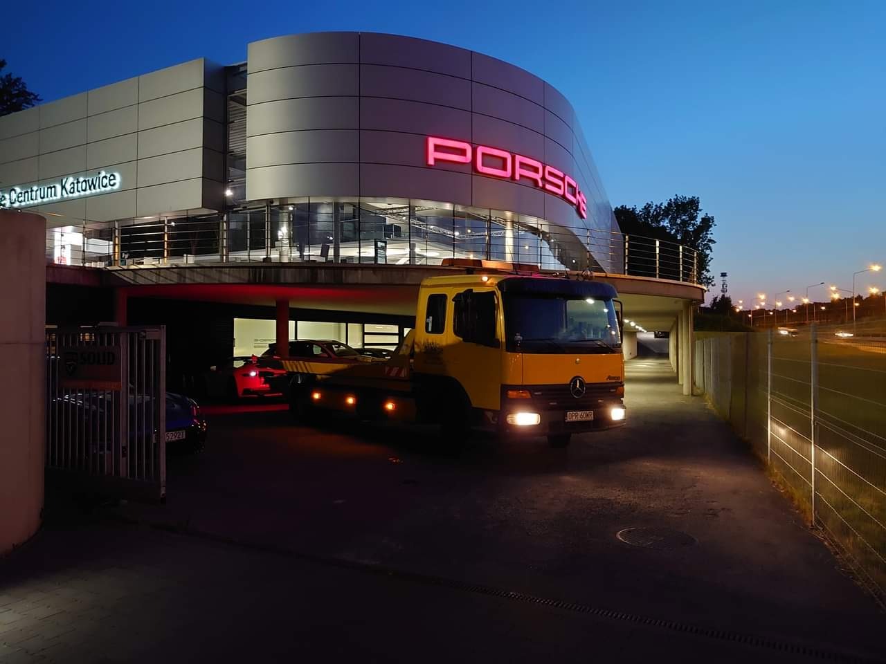 Żółta laweta Mercedes-Benz z włączonymi światłami stoi przed budynkiem Porsche Centrum Katowice, wieczorne oświetlenie.