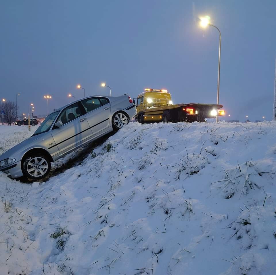 Srebrny samochód osobowy częściowo zjechany z drogi, stojący na zasypanym śniegiem zboczu, holowany przez żółtą lawetę z włączonymi światłami ostrzegawczymi o zmierzchu.
