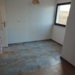 Firma Budowlana SOLARIS - Pokój po częściowym remoncie, widoczne ułożone płytki podłogowe przechodzące w parkiet, ściany pomalowane na jasny kolor, okno z widokiem na budynek.